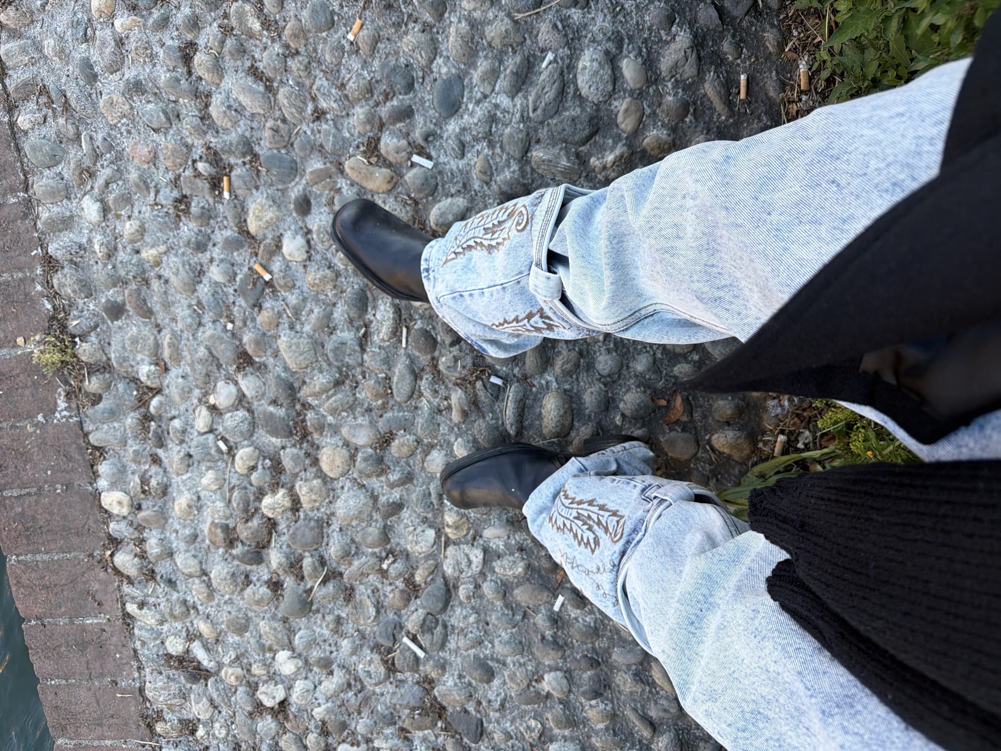 The image depicts a person's lower body and feet standing on a cobblestone surface littered with cigarette butts. The person is wearing light blue denim jeans with an embroidered pattern on the side. They are also wearing black leather boots. A dark-colored garment, possibly a coat or sweater, is visible on the right side of the frame. The background consists of the cobblestone surface with the cigarette butts scattered across it. There is some vegetation visible on the right side of the frame. The scene has been identified as being in Milan, Italy.