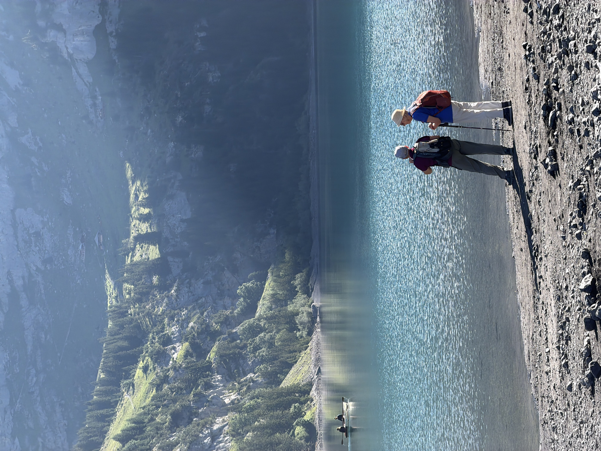 Two individuals, appearing to be hikers, stand on the rocky shore of a calm, turquoise lake. The person on the left wears a burgundy shirt, a grey backpack, and a grey cap. The person on the right wears a blue shirt, a straw hat, a red backpack, and uses a hiking pole. In the distance, a small boat with two occupants navigates the lake. The background features a steep, forested mountainside with scattered buildings and a visible path winding upwards. The scene is illuminated by sunlight, casting shadows on the shore and highlighting the texture of the water. The location is the Oeschinensee lake near Kandersteg, Switzerland.