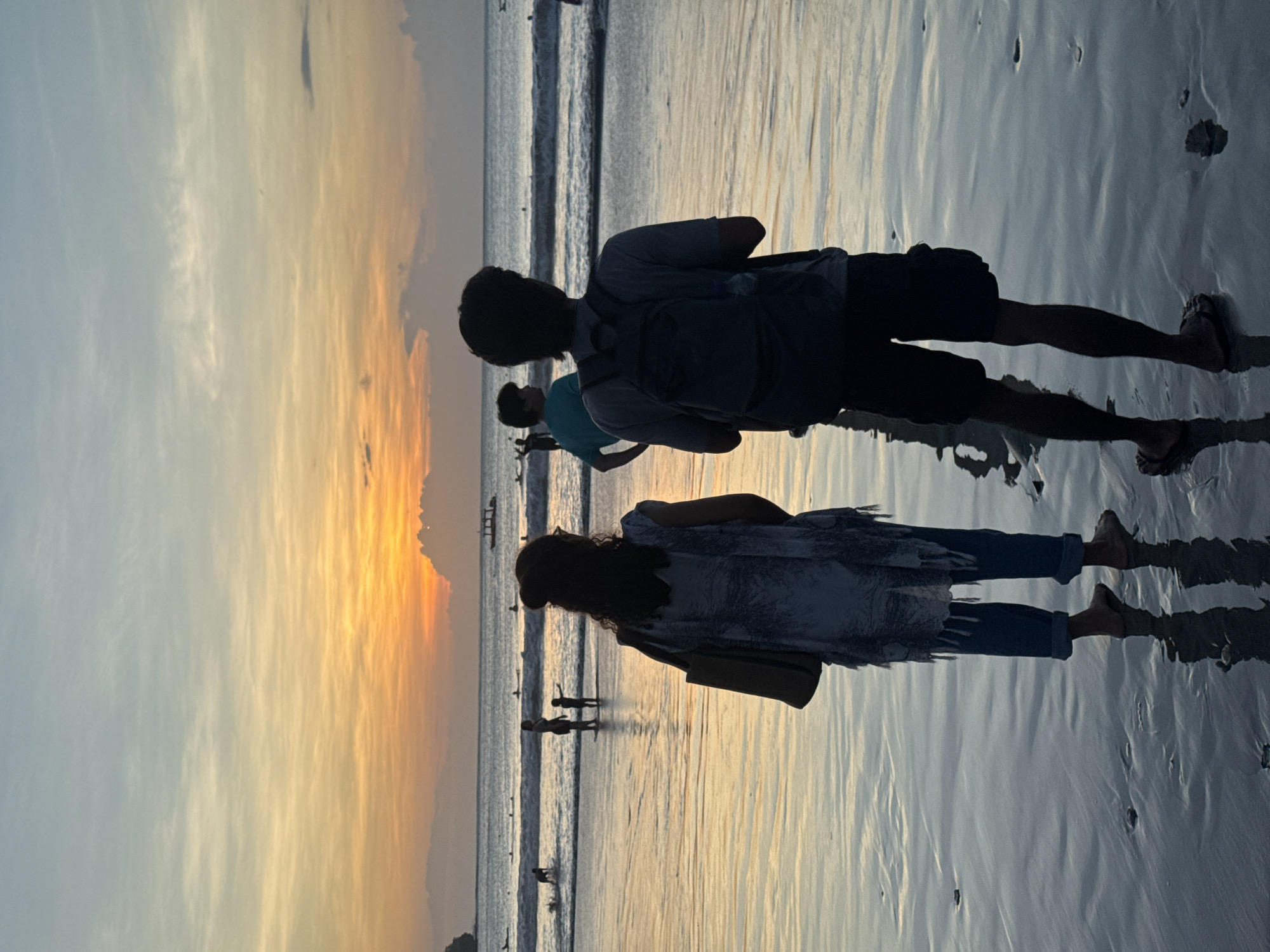 The image depicts a sunset scene at a beach in Kuta Selatan, Indonesia. Silhouetted figures of people are seen walking along the shoreline at dusk. A man and a woman stand in the foreground, with the ocean to the right and the sunset to the left of the frame. Other silhouetted figures are visible further down the beach, along with small boats on the water. The beach surface is wet, reflecting the light of the setting sun.