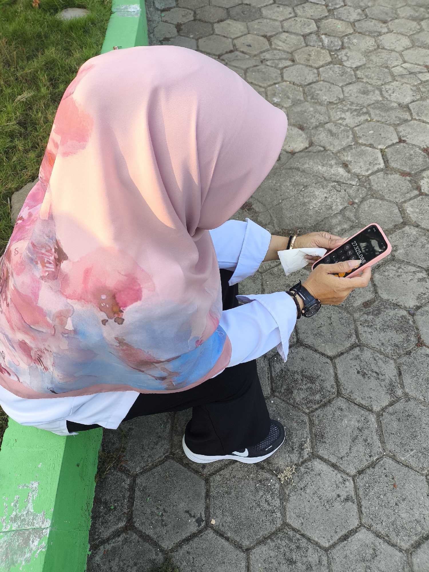 A person wearing a pink hijab with floral patterns, a white long-sleeved shirt, and black pants is sitting on a green curb. The person is holding a pink smartphone in their hands, with a calculator application open displaying "27,325,088". They are also holding a white tissue. A black wristwatch is visible on their left wrist. The background consists of hexagonal concrete paving stones and some grass. The scene is outdoors, likely in a park or public area. The image does not contain enough information to definitively place it in Cilegon, Indonesia.
