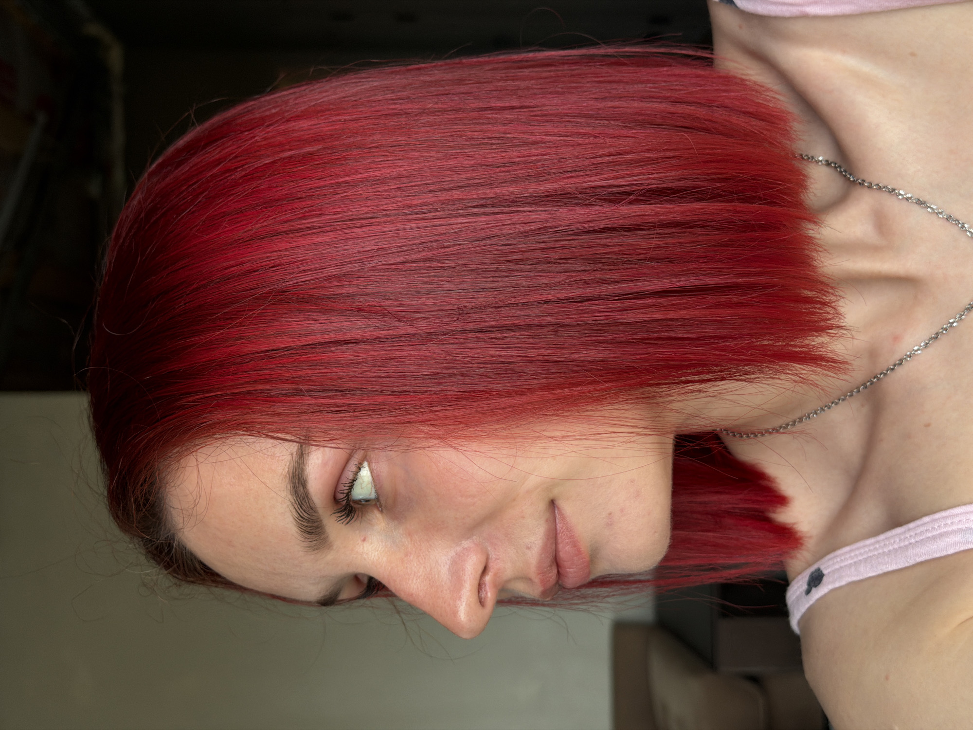 A close-up, rotated portrait of a woman with short, red, chin-length hair, fair skin, and light eyes. She wears a silver chain necklace and a pink top with black polka dots. She is looking down and to the left. The background is a blurred mix of dark and light tones, with indistinct objects suggesting an interior space. There's no indication this photo was taken in Chaikovsky, Russia.