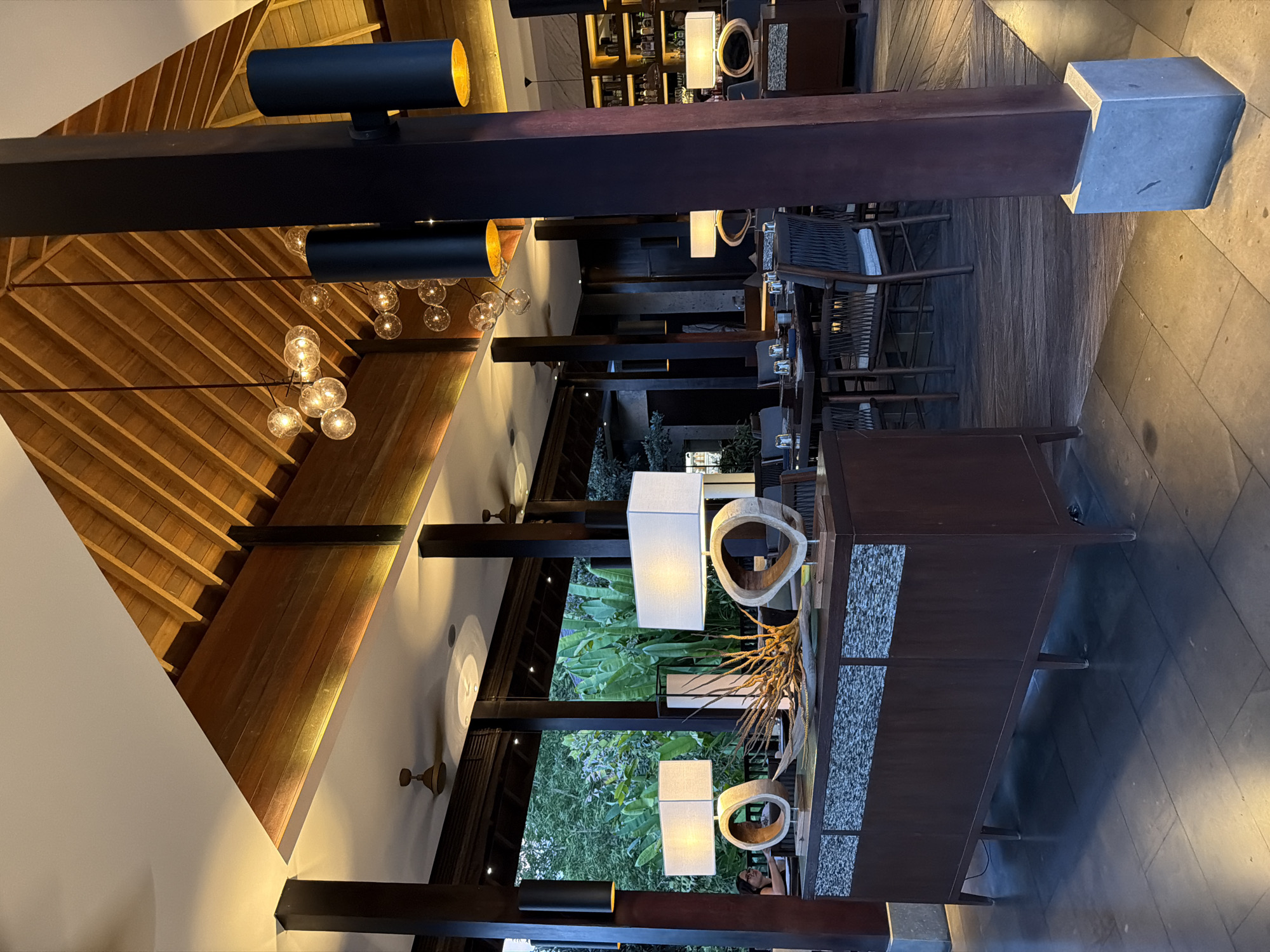 The interior of a restaurant is captured in this photograph. Visible are a wooden, peaked ceiling, dark wooden beams, track lighting, and decorative hanging lights. Tables and chairs are set for dining, and a bar area with shelving is also present. Green foliage is visible through open areas, suggesting an indoor-outdoor setting. The location is identified as Kediri, Indonesia, though specific local details are not visible in the photograph.