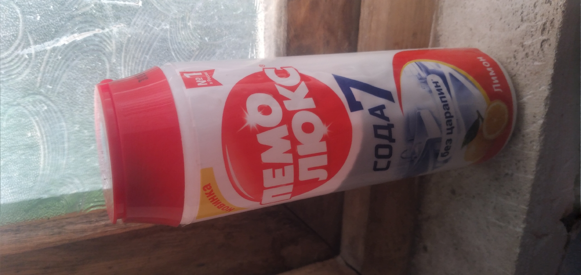 The image shows a white bottle with a red cap, tilted on a windowsill. The bottle has a red label with white text that appears to read "ПЕМО ЛЮКС" and "СОДА 7". There is also text in Russian on the label, including "новинка", "№ 1 в России", "без царапин", and "Лимон". The bottle is positioned in front of a window with a patterned glass. A wooden frame is visible on the left side of the image. The windowsill is made of a rough, grey material. There are two yellow circles with an orange slice and a green leaf graphic on the lower part of the bottle. The lighting suggests it is daytime, with natural light coming from the window. The overall scene depicts a common household product displayed on a windowsill. The location is not specified in the image.