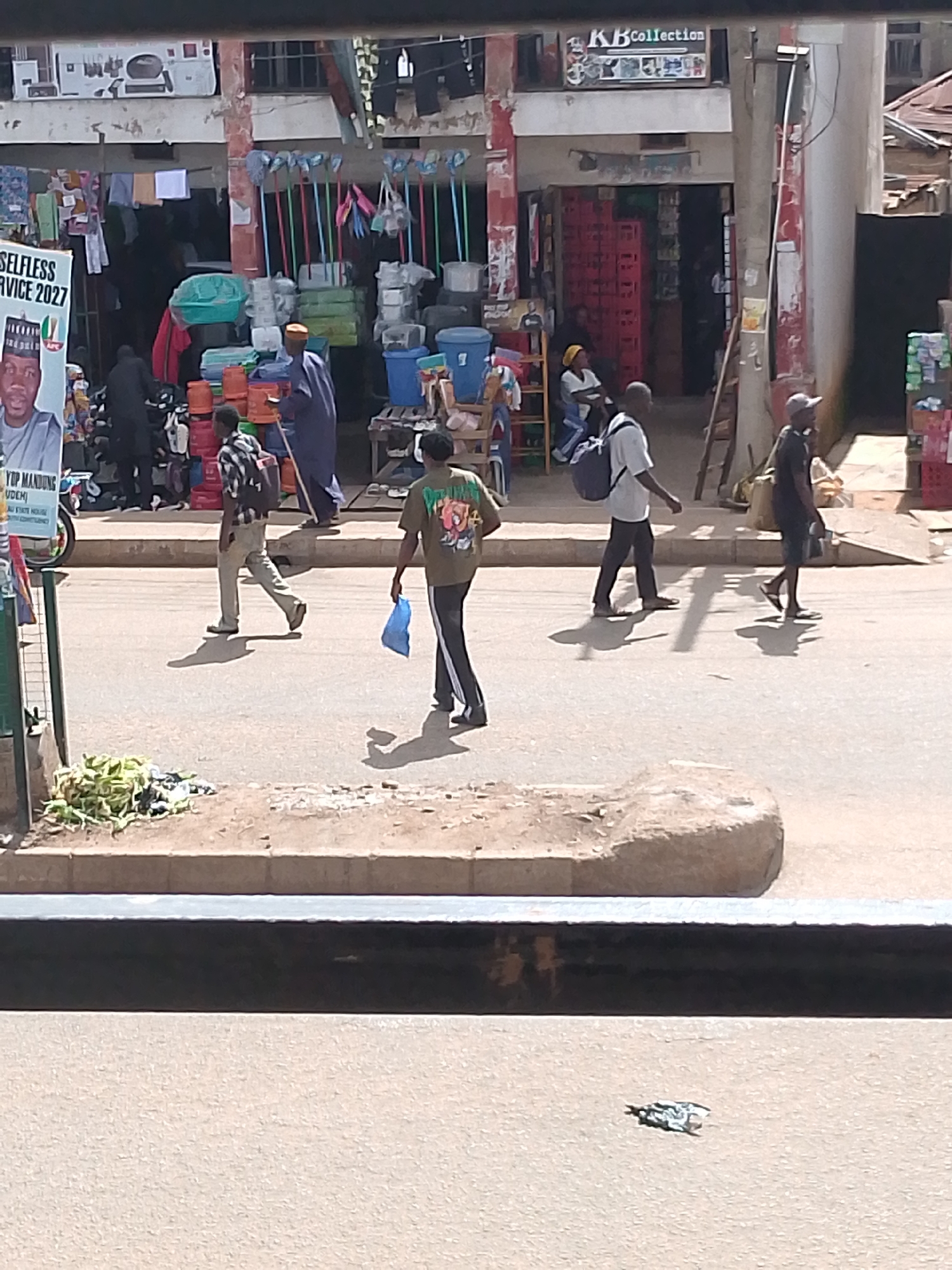 The image depicts a street scene in Bukuru, Nigeria. Several individuals are walking across a paved road. In the background, a market or shop is visible, with stalls displaying various items such as buckets, cleaning supplies, and what appear to be stacks of crates. A poster featuring a man in political attire with the text "SELFLESS SERVICE 2027" is also prominent. The time of day appears to be daytime, with strong shadows cast by the people, suggesting sunny weather. The scene is bustling with activity, typical of a market environment in a populated area. One person is carrying a blue plastic bag, and another is wearing a backpack. The architecture of the buildings in the background suggests a tropical or subtropical region.