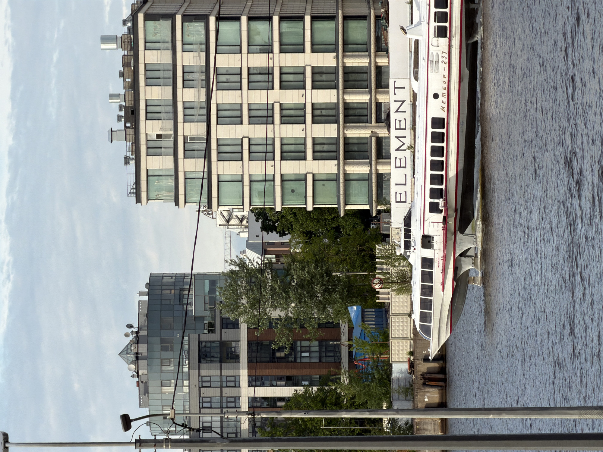 An urban waterside scene unfolds under a cloudy sky, showcasing modern architecture alongside a body of water. In the foreground, metal railings and utility poles with overhead wires frame the view.
To the left, a multi-story building features a facade of light-colored panels and dark-framed windows, with various ventilation units and equipment on its roof. Behind it, another large, contemporary building displays a predominantly glass facade, accented by darker sections and crowned with a distinct pyramidal roof feature. Several trees with green foliage are visible between and in front of these structures, providing a natural contrast.
On the right side of the image, a long, white passenger boat, identified by the large black text "ELEMENT" on its side, is positioned close to a quay. Further along its hull, the text "METEOR-237" indicates it is likely a Meteor-class hydrofoil, a common type of high-speed passenger vessel. The boat has numerous rectangular windows running along its length and a red stripe near its waterline.
The tranquil water surface, likely a river or canal, stretches across the right portion of the frame, exhibiting subtle ripples. Behind the boat and trees, some industrial elements like concrete blocks and a blue tarp-covered object are discernible. The scene captures a daytime moment with diffused light, characteristic of an overcast sky in an urban setting, possibly in Saint Petersburg, Russia, given the context.