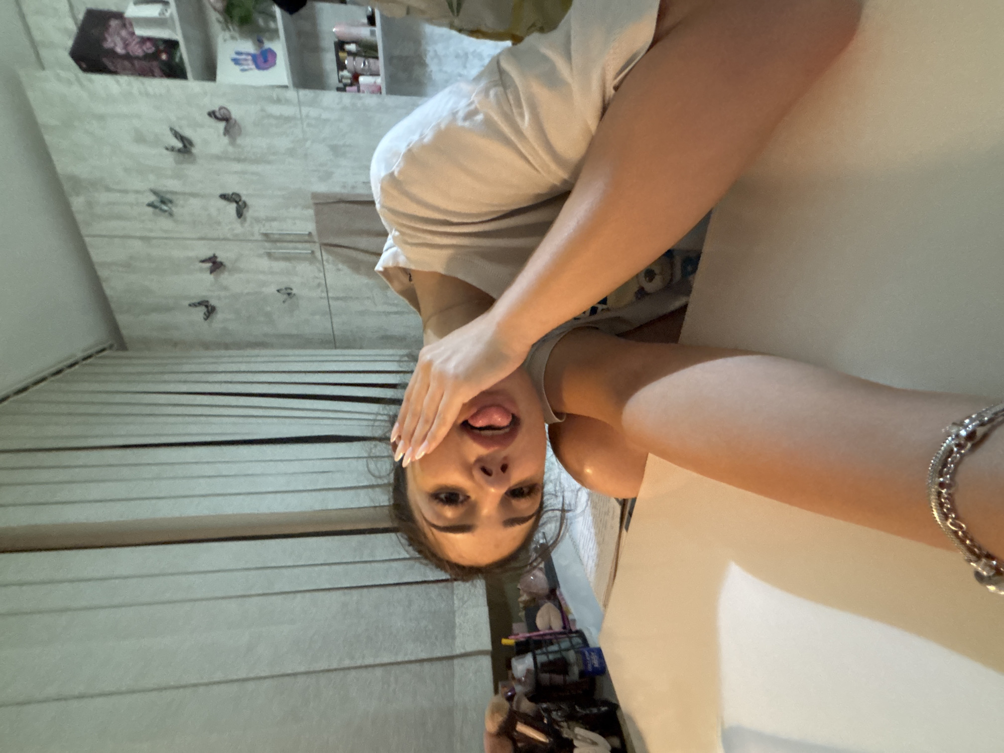A young woman with dark hair is resting her left hand on her face and sticking her tongue out. She is wearing a white t-shirt. She is seated at a table with a white surface. There are blinds on the left side of the image, and a wall with butterfly decals on the right side. A shelf with various items is visible on the far right. The lighting suggests an indoor setting. There is no indication of the image being taken in Plovdiv, Bulgaria.