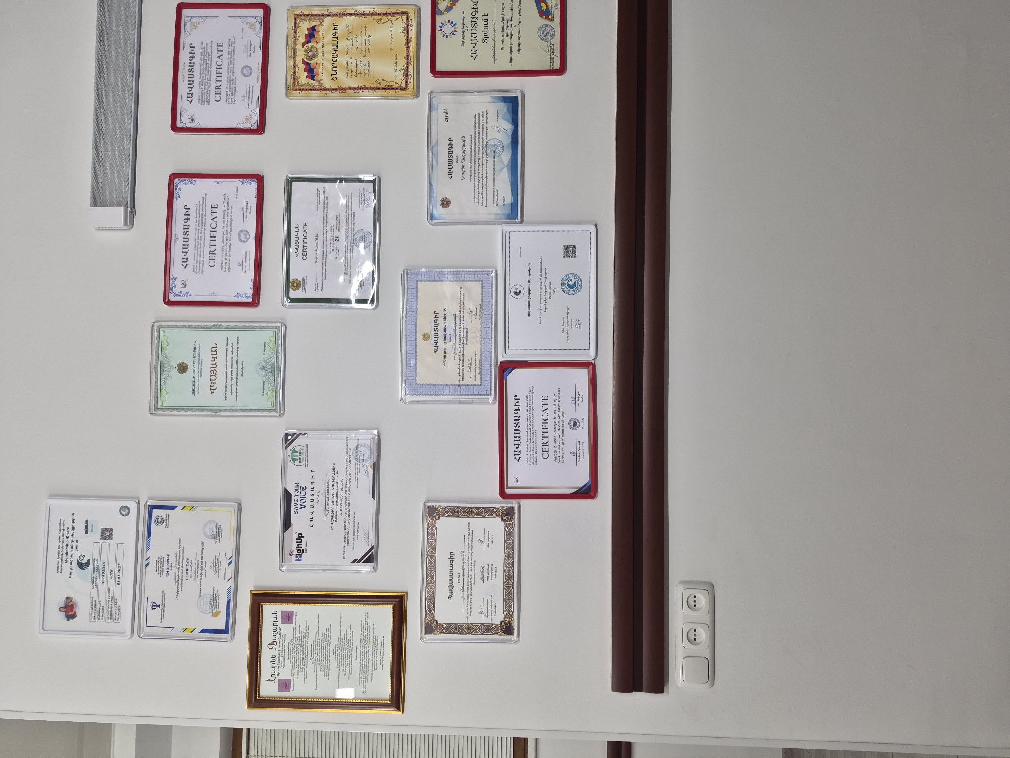 The image captures a wall adorned with a collection of framed certificates, diplomas, and a membership ID card, all displayed in an indoor setting. The wall is light-colored, with a long, horizontal light fixture visible in the upper left and a vertical dark wooden beam running down the right side. Towards the bottom right, a white electrical outlet unit with two sockets and a switch is mounted on the wall.
The numerous documents vary in size and framing, with some in red frames, others in gold, clear plastic, or blue. Visible text indicates many are "ՀԱՎԱՍՏԱԳԻՐ" (Certificate) or "CERTIFICATE". One prominent document is a "Membership ID card" from the "Armenian Speech Therapy Association" with the number ASTA01046, valid until 01.01.2027. It includes a small photo of a person and indicates membership since 2026. Several certificates are awarded to "Lusine Ghazaryan" (Լուսինե Ղազարյանին), with some mentioning training on "Work carried out with children with autism organized by 'Positive Room' psychological center," and another on topics like "Sensory Integration, Behavioral Thread, Group Work." Logos such as "HighUp" and phrases like "SAVE YOUR VOICE" and "ՇՆՈՐՀԱԿԱԼԱԳԻՐ" (Letter of Thanks) are also discernible. Many certificates feature Armenian script and official-looking seals, suggesting professional qualifications and achievements. The display conveys a sense of professional accomplishment and ongoing education, likely belonging to a therapist or psychologist in Yerevan, Armenia.