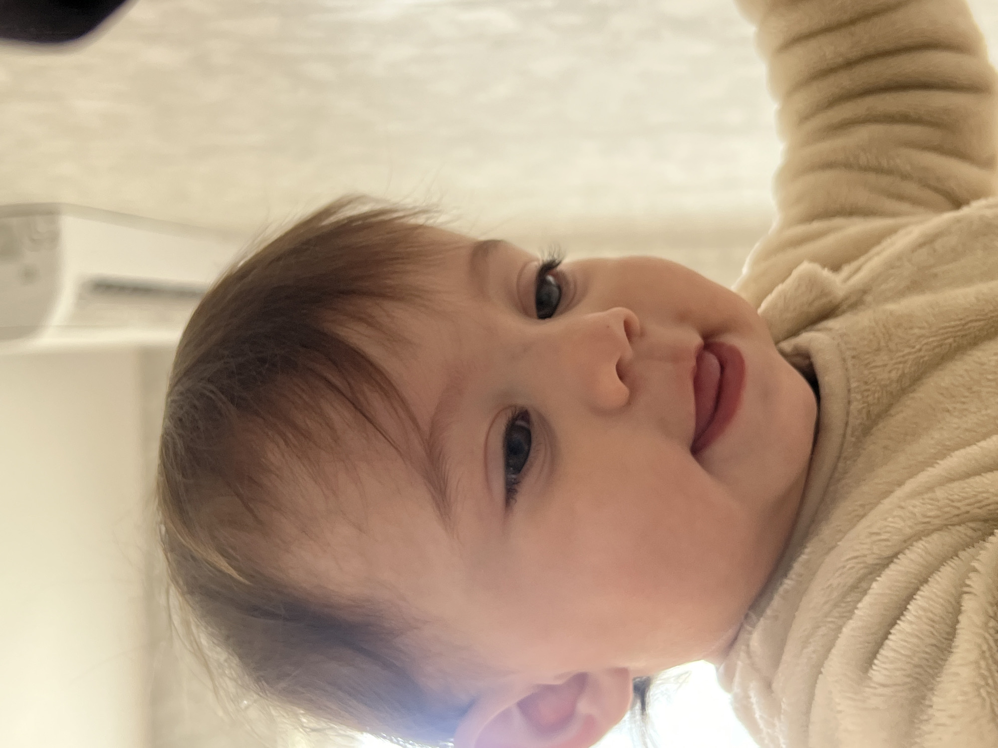 The scene depicts a baby within a room in the city of Elena, Bulgaria. An infant or young toddler with light skin and brown hair, visible from the chest up, is positioned centrally within the frame. The subject's blue eyes are directed downwards and slightly to the right. The mouth is slightly open, revealing the tongue in a relaxed or curious expression. The baby wears a light-colored, possibly cream or beige, fleece garment with a textured surface. A portion of the baby's right arm, covered by the same garment, is visible on the right side of the frame. The background is out of focus, appearing as a light-toned interior wall. A white, rectangular wall-mounted air conditioning unit is partially visible in the upper-left background.