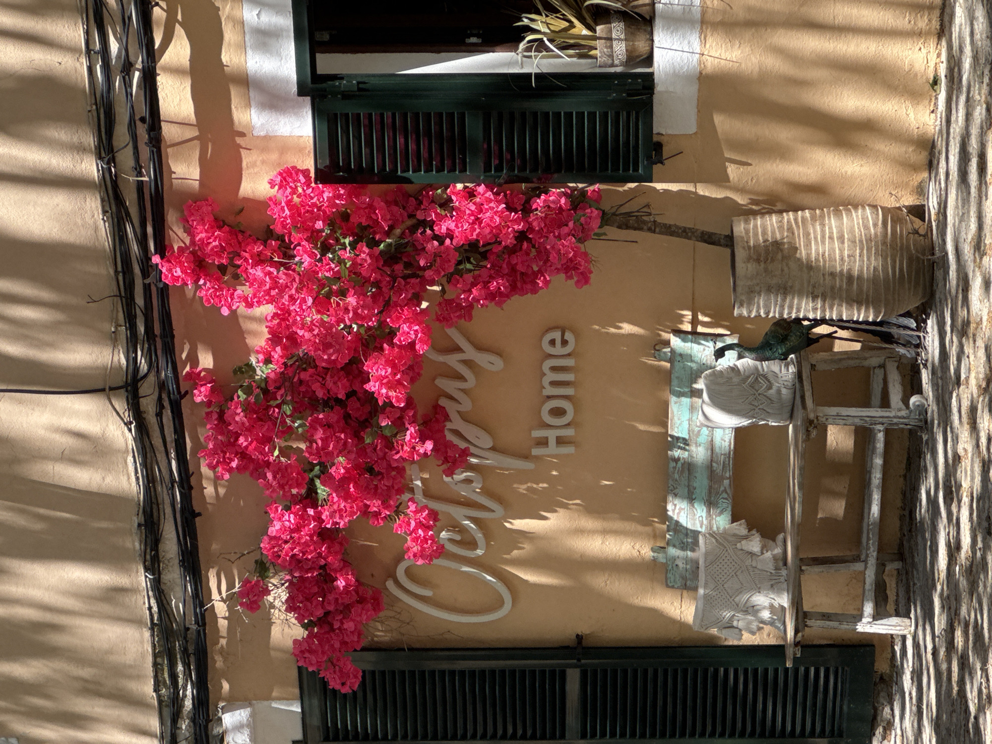 The image shows the exterior of a building in Valldemossa, Spain. The wall is painted a muted yellow. A vibrant pink bougainvillea covers part of the wall and a window with green shutters. There is a white sign that reads "Octopus Home." A white wooden chair with pillows sits on the wall. A window with green shutters is located above the bougainvillea. Electrical cables run along the left side of the wall. Shadows from nearby trees fall across the scene.