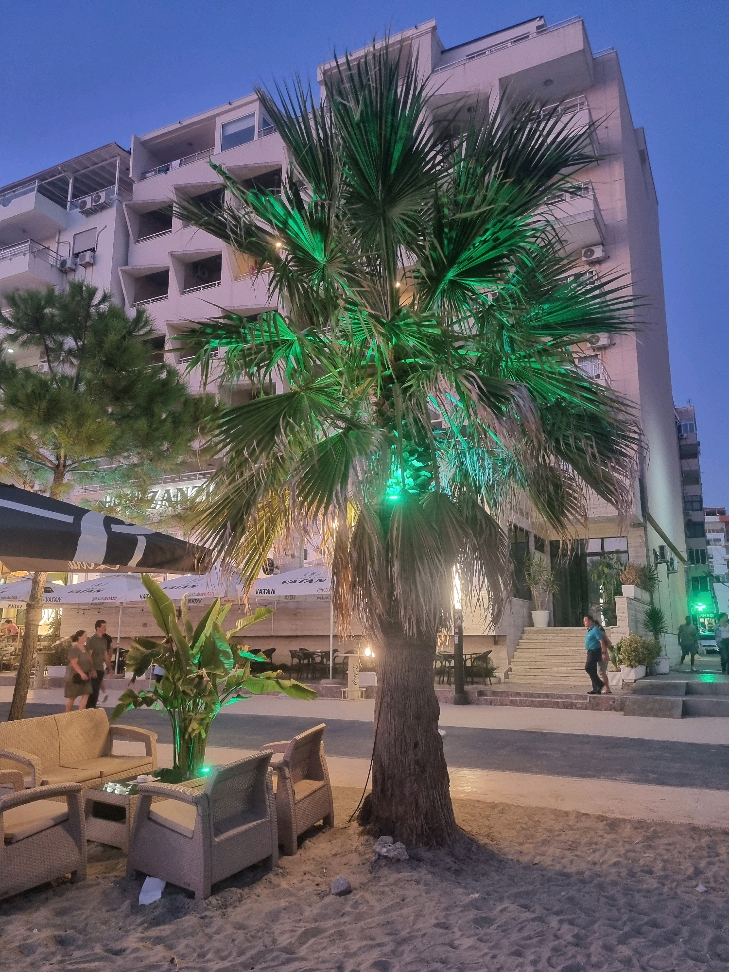In Bashkia Durrës, Albania, at dusk, a palm tree with green lights illuminating its fronds stands prominently on a sandy area. Behind the palm tree, a modern building with multiple floors and balconies is visible. The building appears to be a hotel or an apartment complex. To the left of the palm tree, there are several white umbrellas and outdoor seating areas with rattan furniture, suggesting a restaurant or cafe. People are seen walking in the background. The sky is a deep blue, indicating the transition from day to night.