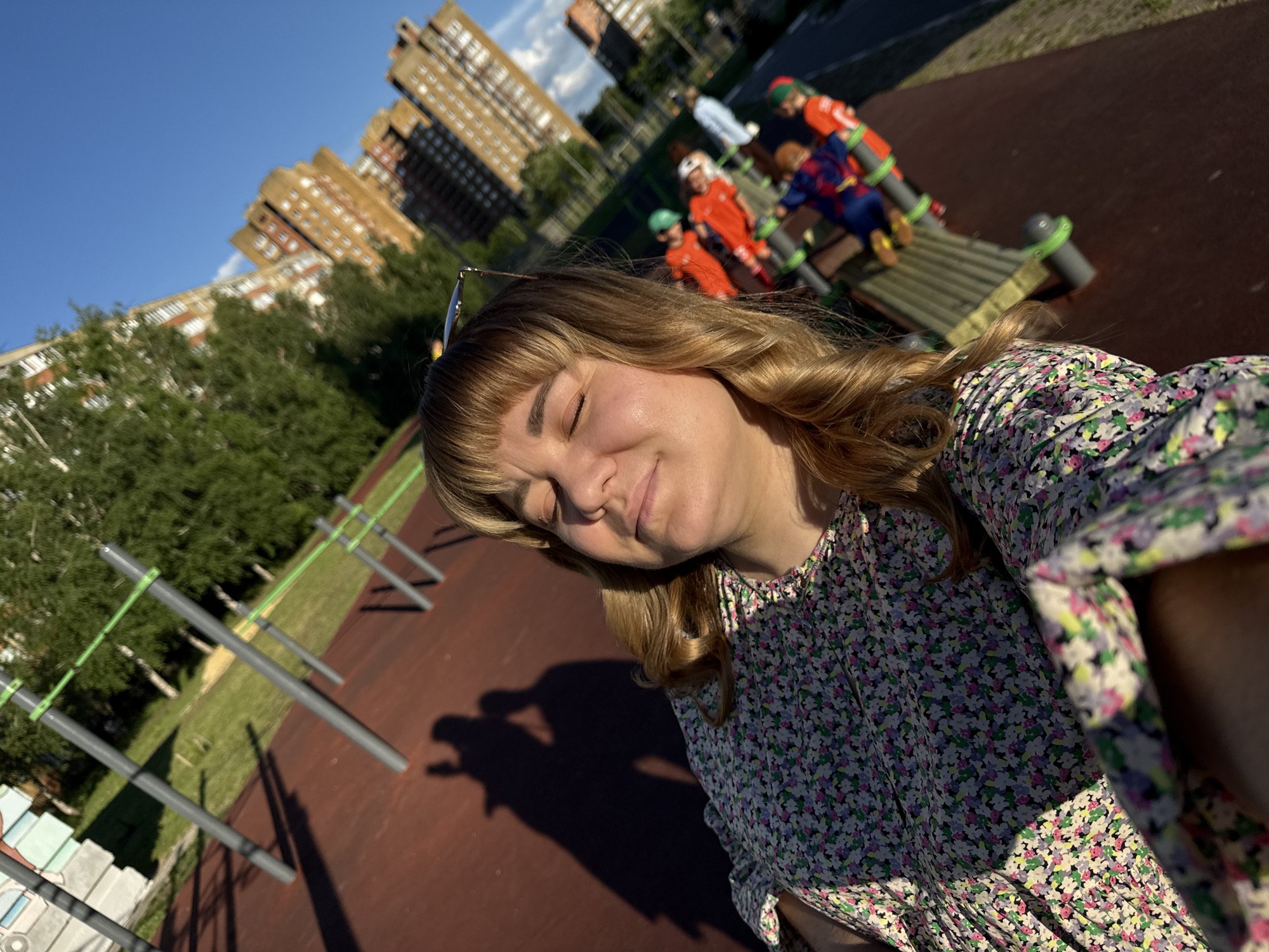 The image captures a selfie of a young woman with blonde hair and bangs, wearing a floral dress. She has her eyes closed, a slight smile on her face, and sunglasses perched on her head. The setting appears to be an outdoor playground or sports area. In the background, several children are visible on a multi-level playground structure. Some children are dressed in brightly colored athletic wear, one in what looks like a superhero costume, and another in an orange outfit with green accents.
The playground features red rubberized surfacing and metal bars, possibly for exercise or climbing. Beyond the playground, there are green trees and tall apartment buildings, suggesting an urban environment. The sky is a clear, bright blue, indicating fair weather and likely daytime. The angle of the sunlight and the long shadows cast on the ground suggest it is late afternoon. The presence of the apartment buildings and the overall aesthetic might point to a location in Stary Oskol, Russia, as suggested. The woman's relaxed expression and the sunny atmosphere convey a sense of peace and enjoyment. There is no visible text in the image.