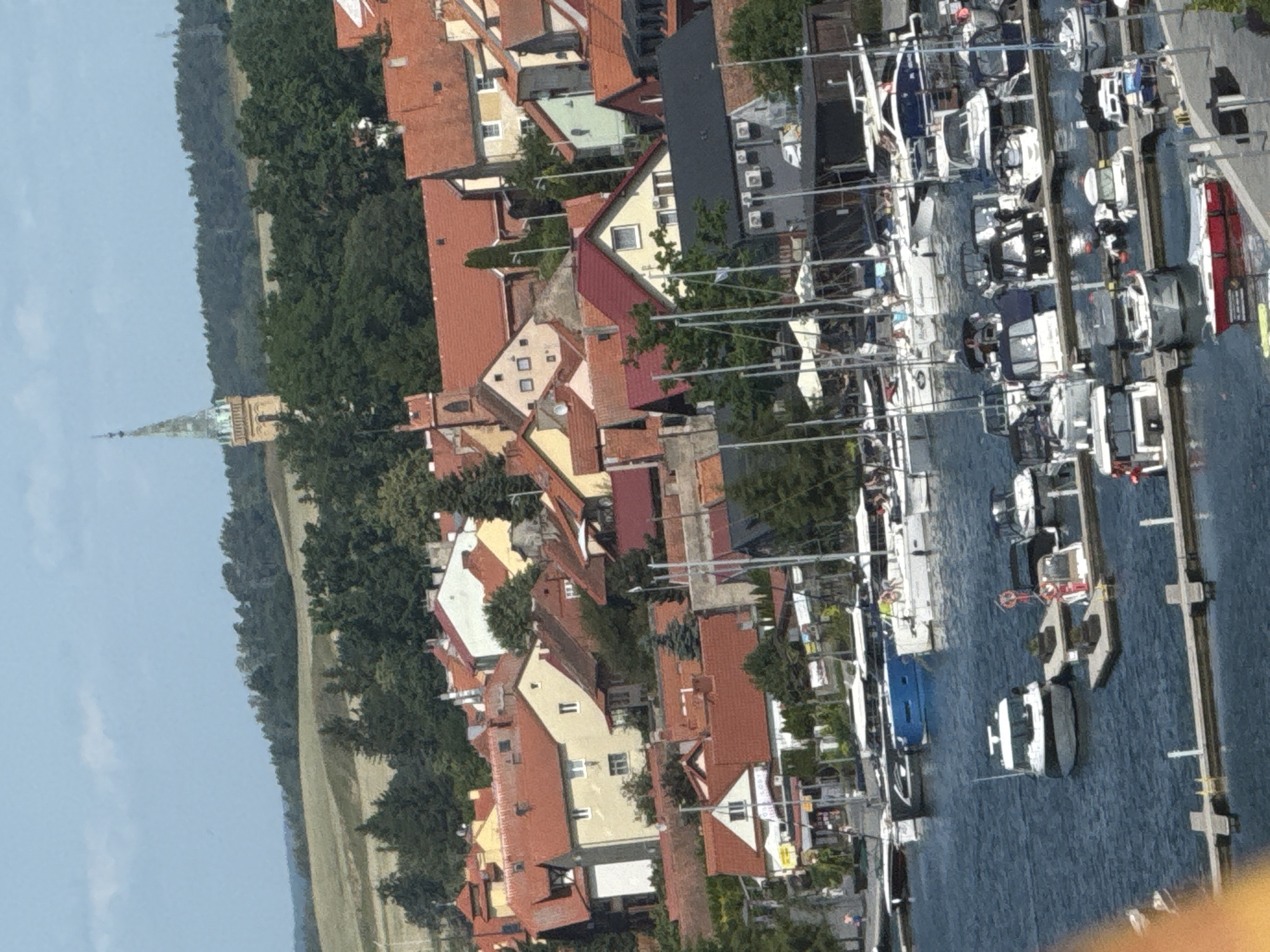The image depicts a panoramic view of a waterfront town in Mikołajki, Poland, likely during daytime under a cloudy sky. The scene is dominated by a harbor filled with numerous boats and yachts of various sizes, some docked and others moored. Adjacent to the harbor, a cluster of red-tiled roof buildings forms the town's skyline. In the background, rolling hills covered with greenery and trees are visible, with a prominent church spire rising above the foliage in the distance. The overall impression is of a scenic, bustling European lakeside town.