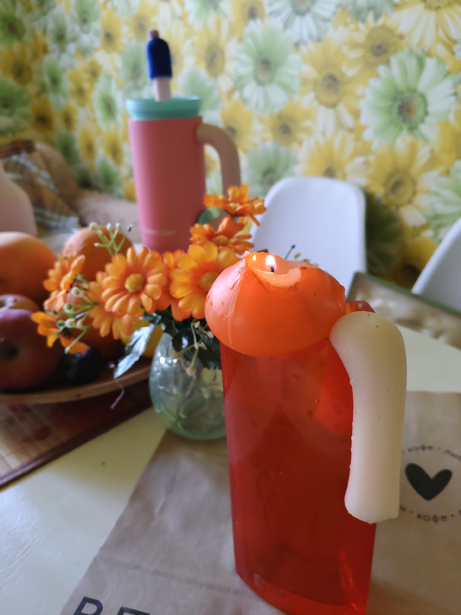 A candle in the shape of a mug, with a white handle, burns on a table. The candle is orange and has a lit wick. In the background, there are two mugs, one pink and one with a blue lid, along with a vase of orange flowers, fruit on a wooden plate, and a bag with a heart emblem. The scene is set against a floral wallpaper. The location is a domestic setting in Rostov-on-Don, Russia.