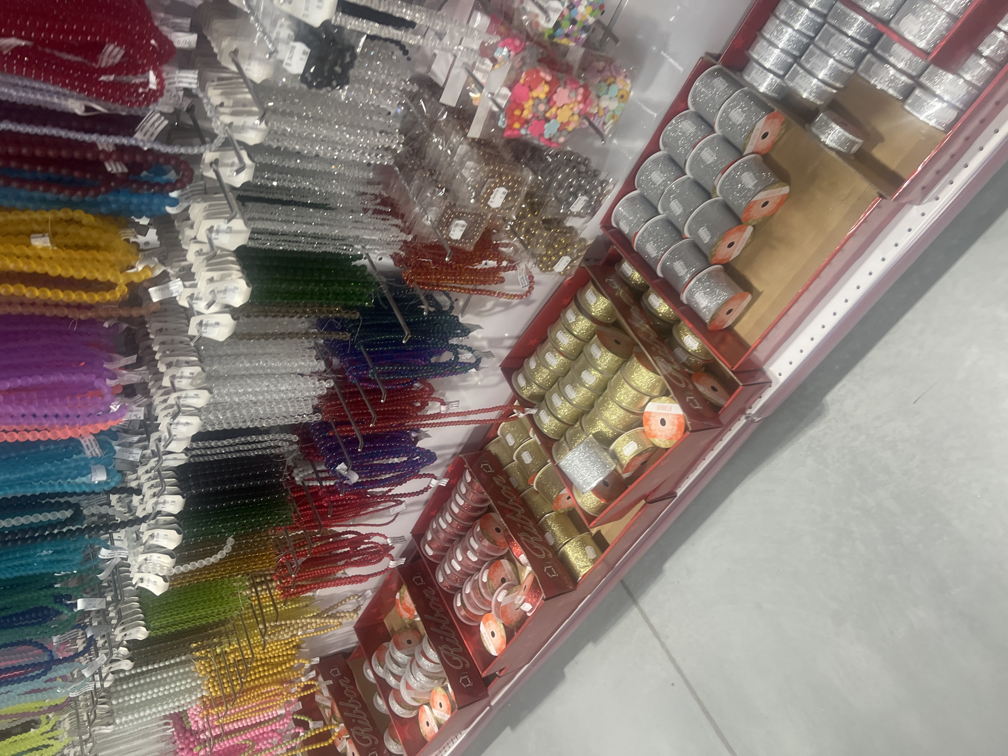 The image shows a display of crafting supplies in what appears to be a retail store. Strands of colorful beads, arranged by color, hang on the left. To the right are shelves stocked with rolls of ribbon in various colors and textures, including silver, gold, and red. The scene has a slightly tilted perspective. There is no information in the image about its location in Burgas, Bulgaria.