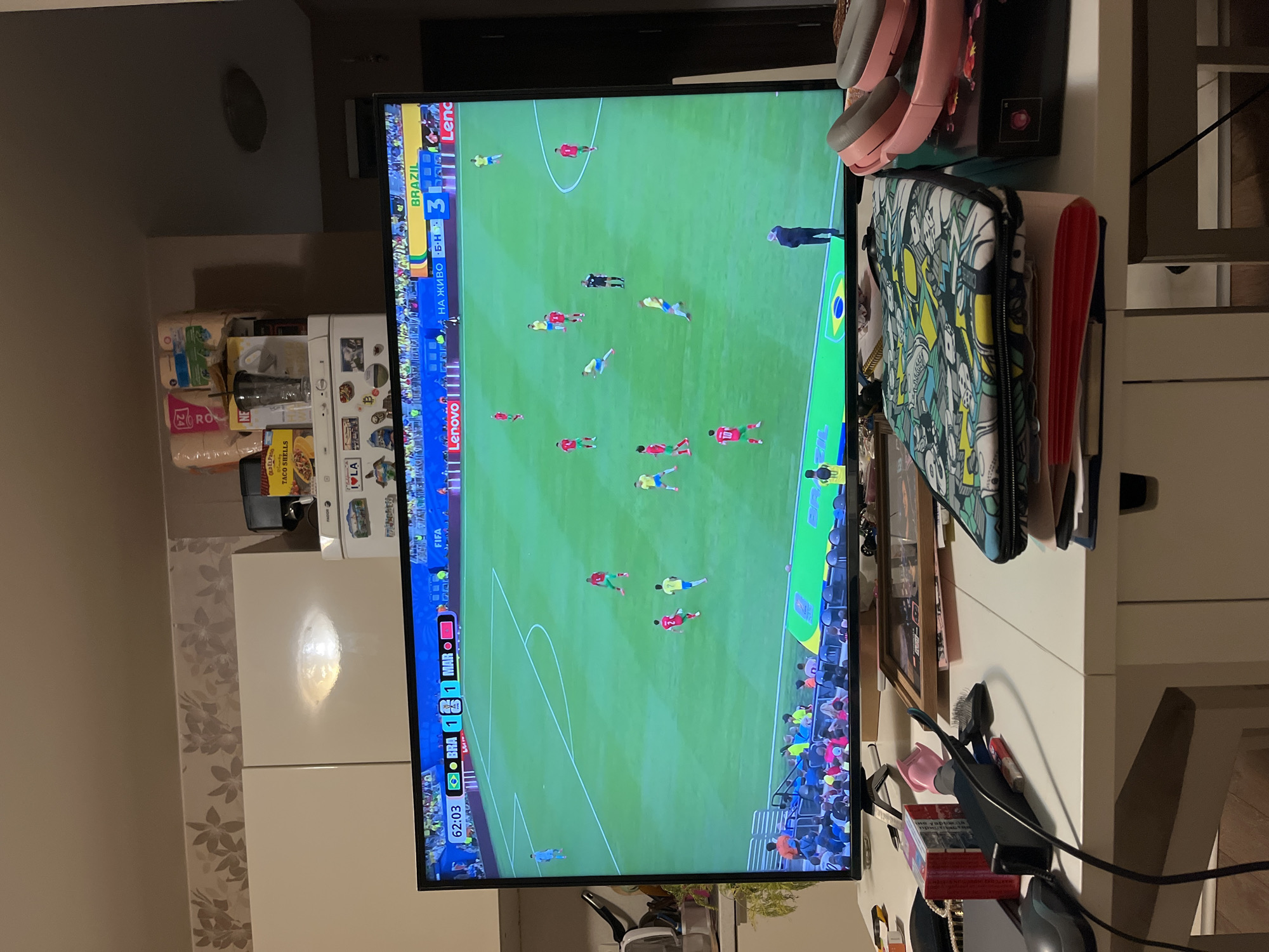 The image shows a television screen displaying a soccer match. The score on the screen shows Brazil leading 3-1 against Morocco, with the game time at 62:03. The match appears to be taking place on a bright, green field. In the foreground, a white table is visible with various objects, including a laptop sleeve with a colorful, cartoonish print, a pair of pink headphones, a framed picture, a black computer mouse, and a red object that resembles a stapler. The background suggests an indoor setting with a wall decorated with floral wallpaper and white cabinets. The lighting in the room is artificial, casting a glow on the wall. Based on the visible elements, the location is likely a living room or common area within a residential apartment in Sofia, Bulgaria. There are no people directly depicted in the foreground, but figures are visible on the television screen playing the soccer match. The overall scene is domestic and casual, with the television being the focal point of interest.