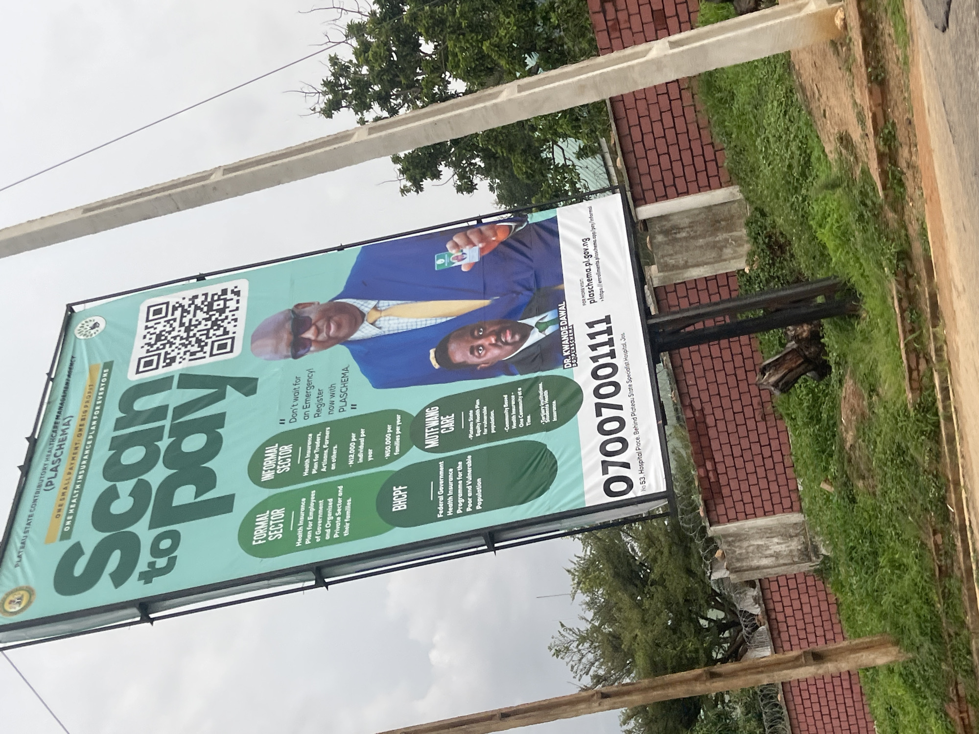 This is a vertical, eye-level outdoor shot of a large billboard in Jos, Nigeria. The billboard advertises the Plateau State Contributory Healthcare Management Agency (PLASCHEMA) and its health insurance plans. The main subjects are two men in business attire, one in a blue suit and the other in a dark suit, both smiling and appearing professional. The billboard's dominant colors are teal and white, with large green text that reads "Scan to Pay."
The billboard highlights different health insurance plans: "Formal Sector" for government and private sector employees, "Informal Sector" for traders, artisans, and farmers, and "BHCPF" (Basic Health Care Provision Fund) which is described as a Federal Government program for the poor and vulnerable. It also mentions "MUTFWANG CARE" which is the "Plateau State Equity Health Plan" for vulnerable populations.
Notable details on the billboard include a QR code, a phone number "07007001111," a website address "plasechema.pl.gov.ng," and a physical address "No 53, Hospital Place, Behind Plateau State Specialist Hospital, Jos." The text indicates that it's "One Small Payment, One Big Profit" and "One Health Insurance Plan for Everyone." The image appears to be taken during the day, with an overcast sky suggesting cloudy weather. The setting is outdoors, with a brick building and lush green vegetation visible in the background, along with a power line crossing the sky. The overall impression is one of a public service announcement promoting accessible healthcare.