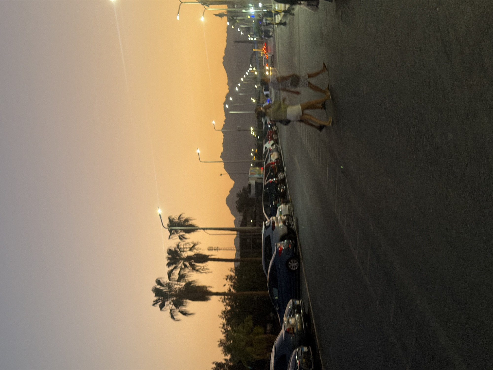 The image depicts a street scene in Sharm El Sheikh, Egypt, during dusk. The sky is a gradient of soft orange and pale blue, typical of a sunset. Several palm trees line the left side of the street, with a row of parked cars to their right. The cars are of various colors and models.
Further down the street, streetlights are illuminated, casting a warm glow. In the mid-ground, three individuals are crossing the street. The two in the foreground are women, one wearing a light-colored skirt and top, and the other a striped dress. They appear to be walking together. A third person, partially obscured, is walking behind them.
In the background, a mountain range is visible against the twilight sky. The overall impression is one of a relaxed evening scene in a coastal city.