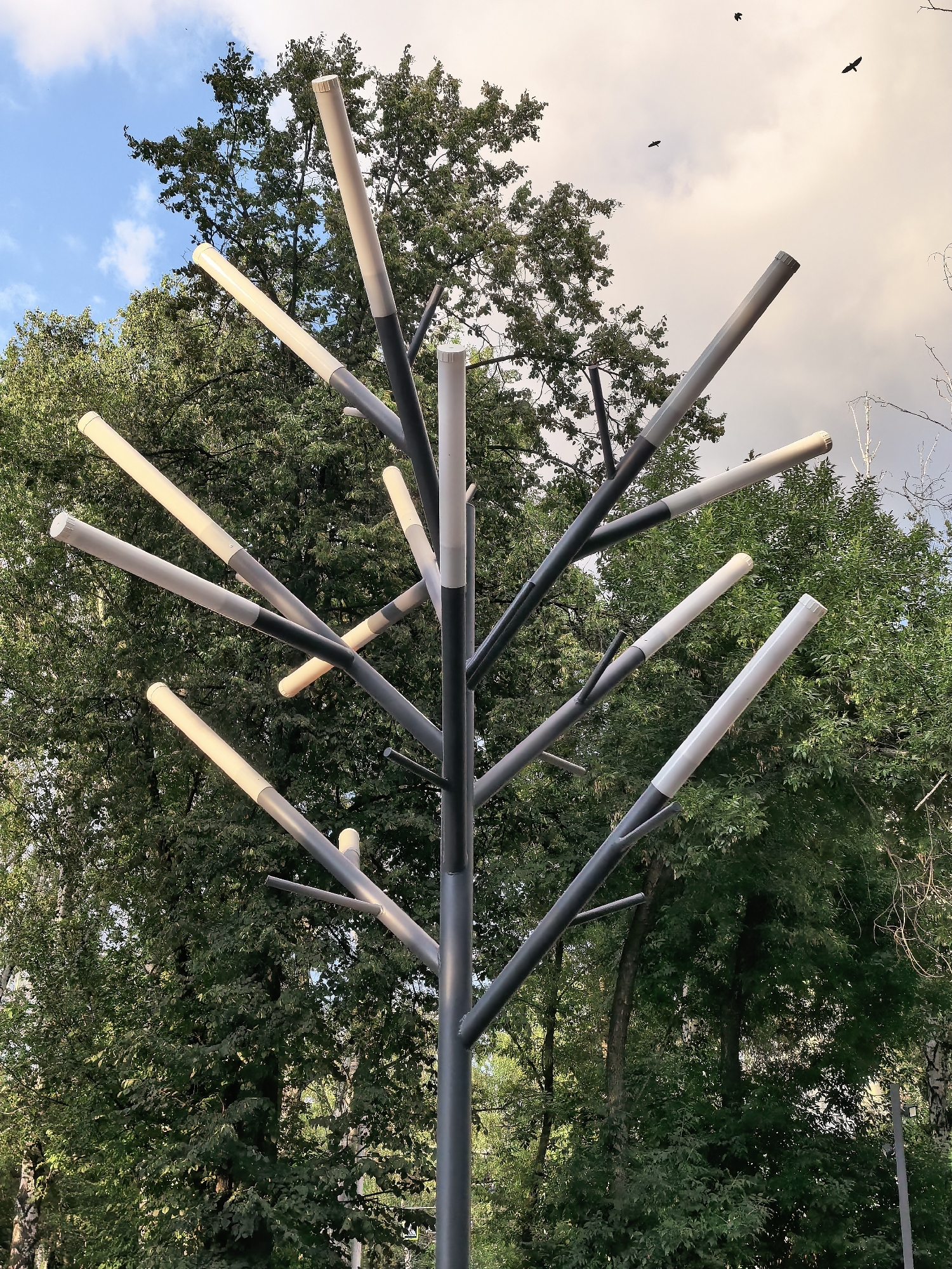 The image shows a modern, tree-like streetlamp with multiple luminous tubes extending from a central pole. The lamp is set against a backdrop of lush green trees and a partly cloudy sky. Three black birds are visible in flight against the sky in the upper right portion of the image. The streetlamp's design is abstract, with its branches reaching outwards at various angles, each capped with a white or beige tube that appears to be illuminated. The foreground is dominated by the metallic structure of the lamp, with its dark gray pole and branches. The background consists of dense foliage from deciduous trees. The lighting suggests it is either early morning or late afternoon. No people or vehicles are visible in the scene. The overall impression is one of a park or public space within an urban setting.
The image is an exterior shot of a streetlamp in the city of Sterlitamak, Russia.