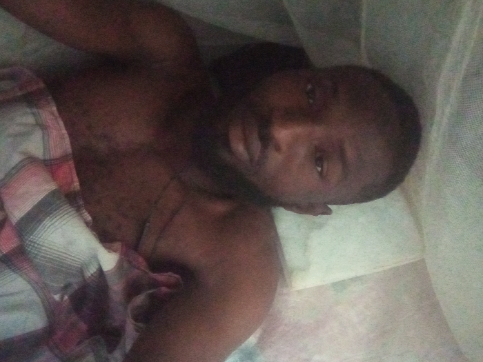A shirtless, dark-skinned man with a beard lies on a bed beneath a mosquito net. His head rests on a white pillow. He is covered partially with a plaid blanket. His eyes are open. He looks at the camera. The lighting is dim. The scene seems to be set indoors, presumably in a bedroom.