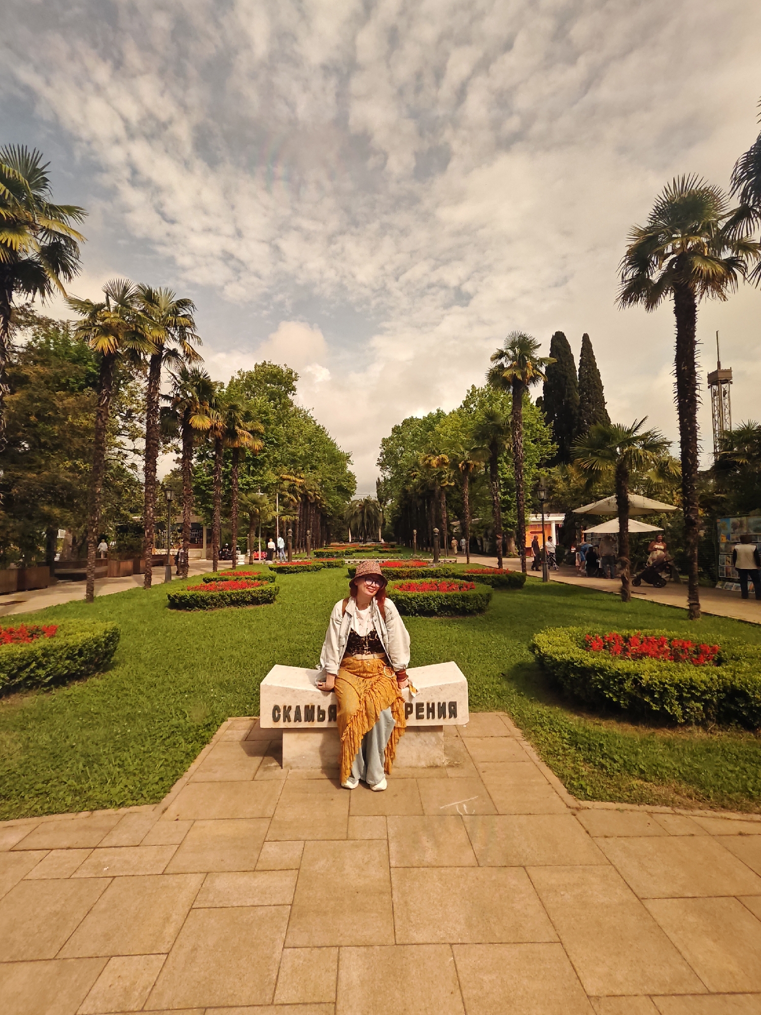 A woman sits on a white concrete bench in a park-like setting. She wears a hat, glasses, a jacket, and an orange fringed skirt or pants. The bench has the word "СКАМЬЯ" inscribed on it, likely meaning bench.
Behind her is a pathway lined with palm trees, flower beds filled with red flowers, and trimmed green bushes. People are walking in the background.
The location is in Sochi, Russia.