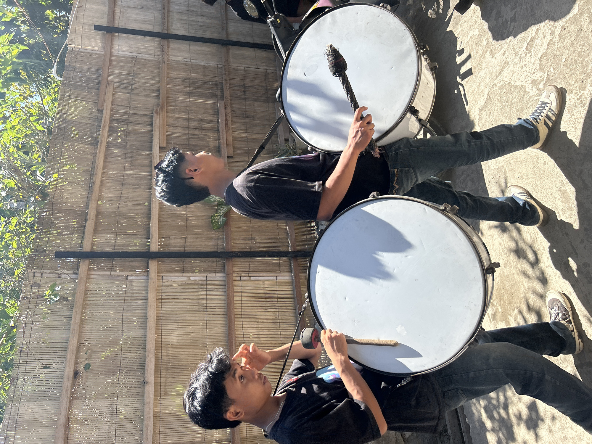Two young males are standing outdoors, each holding a large bass drum. They are positioned on a concrete surface in front of a bamboo fence. The male on the left is wearing a black t-shirt and blue jeans, with a strap around his shoulder supporting his drum. He is holding a drumstick in his right hand, with his left hand near his forehead. The male on the right is also wearing a black t-shirt and blue jeans, and his drum is larger than the one on the left. He is holding a drumstick with a padded end in his right hand, preparing to strike the drum. Both individuals appear to be participating in a musical activity, possibly a parade or performance. The background shows a bamboo fence with wooden posts and foliage from trees overhead, suggesting an outdoor setting possibly in a residential area or public space. Sunlight casts shadows on the ground, indicating it is daytime. There is a hint of a motorcycle to the right of the second male. The location is identified as Kuta, Indonesia.