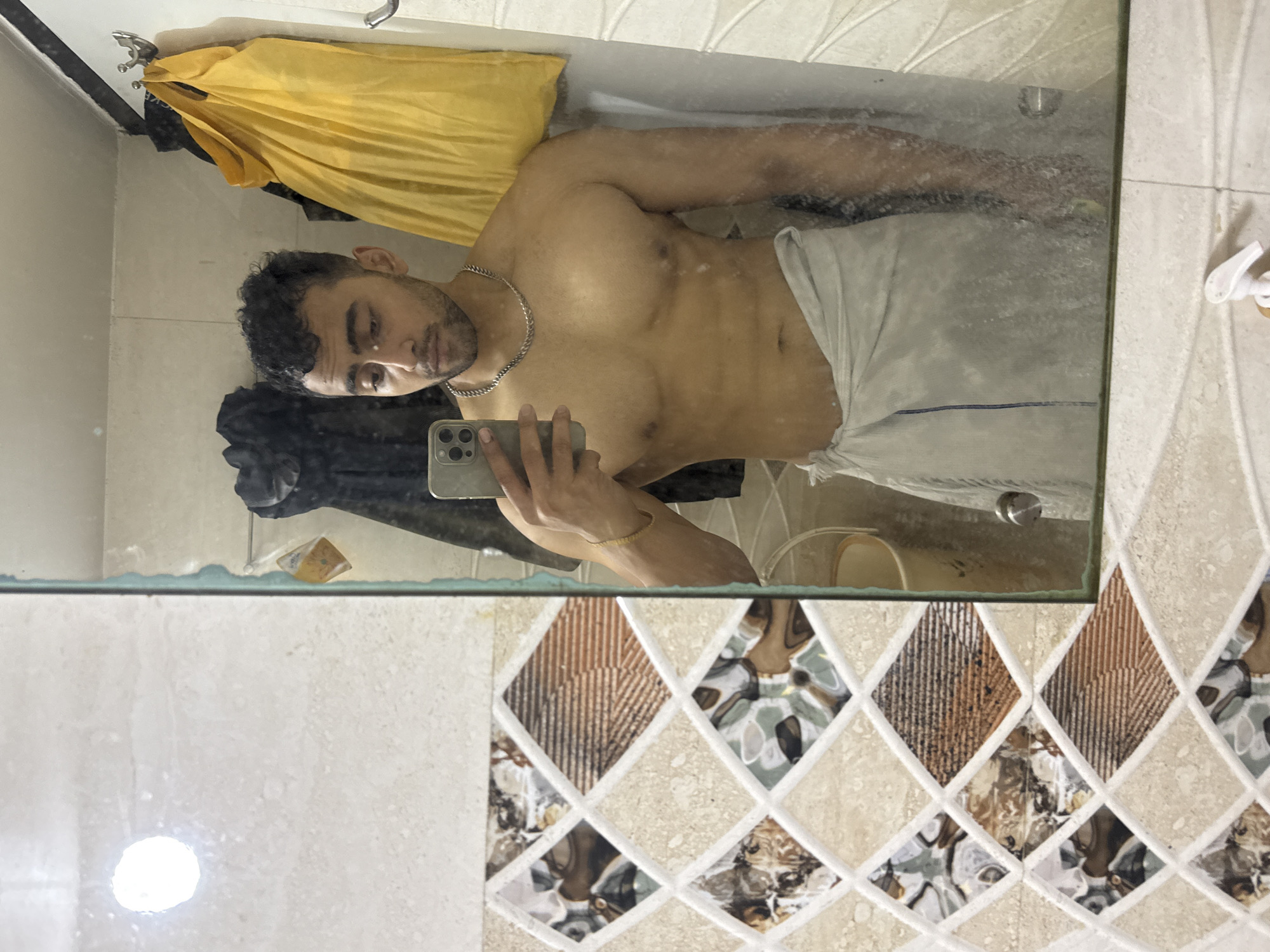 A shirtless man with a towel around his waist is taking a selfie in a bathroom mirror. The bathroom has tiled walls with a patterned design. A yellow garment and other items are visible in the background, hanging near a sink. The location is likely in Kolkata, India, given the man's appearance and the overall style of the bathroom.