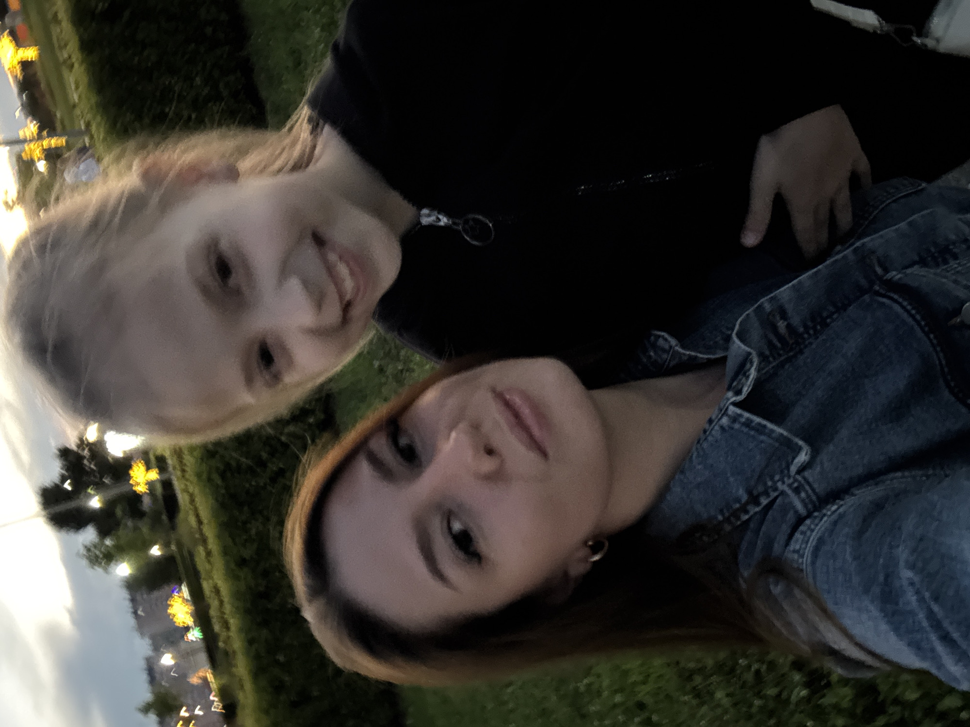 Two females are captured in a selfie outdoors in the city of Unknown, Unknown. The female in the foreground, on the left, has long brown hair and is wearing a denim jacket. The female in the background, on the right, has blonde hair and is wearing a black hoodie. The lighting suggests it is dusk or evening. In the background, there is a park with green bushes and some illuminated decorations or lights. The overall scene appears casual and friendly.