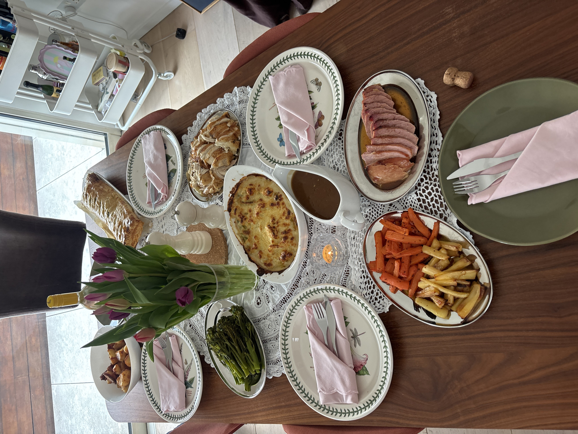 The scene presents a dining table laden with various dishes, likely set for a meal. Dishes include sliced meat, roasted vegetables (carrots and potatoes), scalloped potatoes, asparagus, and bread. Each place setting consists of a plate with a pink napkin and cutlery. A centerpiece features a bouquet of purple tulips and a wine bottle. A white lace tablecloth covers the wooden table, and a wine cork is visible on the right. In the background, a white bar cart can be seen. The setting suggests an indoor dining area, but there is no information suggesting the location in the city of London, United Kingdom. The setting is private and there are no people in the shot.