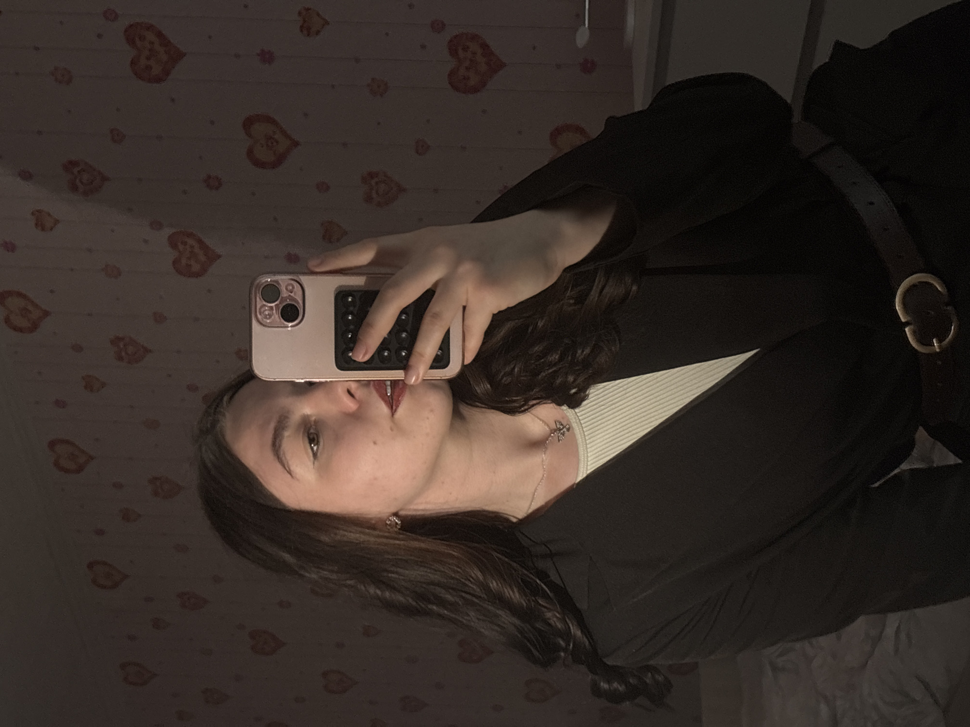 A woman with dark hair is holding a pink smartphone with a black grip accessory near her face. She appears to be taking a selfie. The background features a wall covered in a pattern of stylized hearts. She is wearing a black jacket over what appears to be a white or light-colored shirt. A thin necklace with a pendant is visible on her neck. A dark belt with a gold buckle is visible around her waist. The location cannot be determined solely from the image. The city of Perm, Russia, cannot be determined from the image.