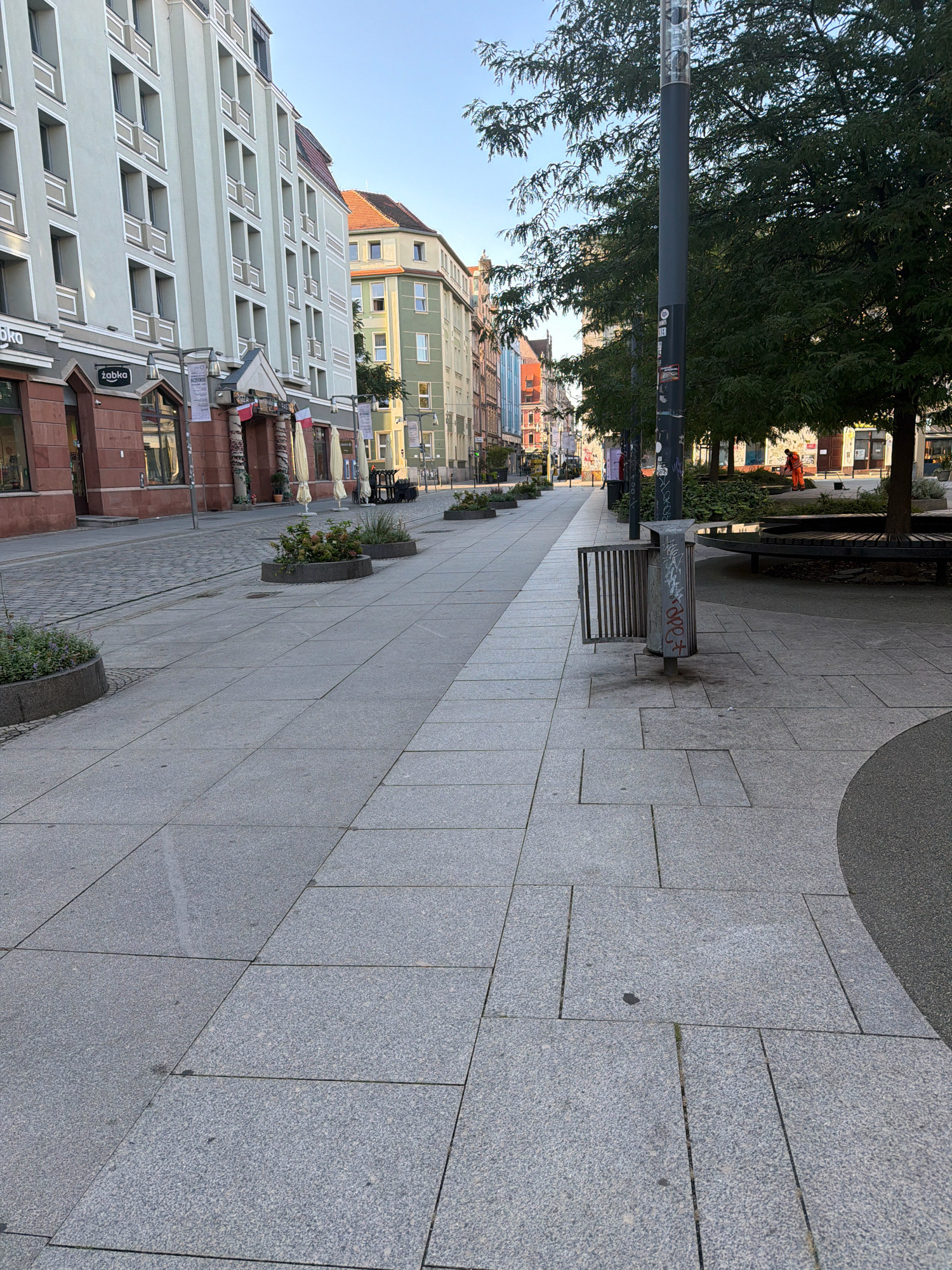 The image depicts a wide pedestrian walkway paved with large, light grey granite tiles, leading into a street lined with multi-story buildings. On the left, a pale green building with rows of rectangular windows dominates the foreground, with red-brick storefronts at street level. A "Żabka" convenience store is visible. On the right, a large tree provides shade over a circular wooden bench and a grey metal trash receptacle. Further down the street, a person in an orange uniform is seen performing groundskeeping duties with a broom. The architecture suggests a European city setting, and the presence of a "Żabka" convenience store specifically indicates the location is Wrocław, Poland. The sky is clear and blue, indicating fair weather.