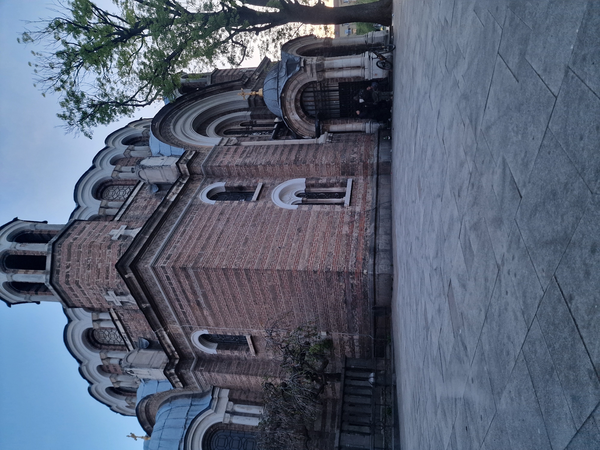This image shows the Saint George Rotunda Church, an early Christian church in Sofia, Bulgaria. The building is constructed of red brick with white arched details. It has a domed roof, and there are trees overhead. On the right side of the image, there is a stone-paved area where a bicycle is parked, and some people are standing. The overall impression is of a historic building in an urban setting at twilight.