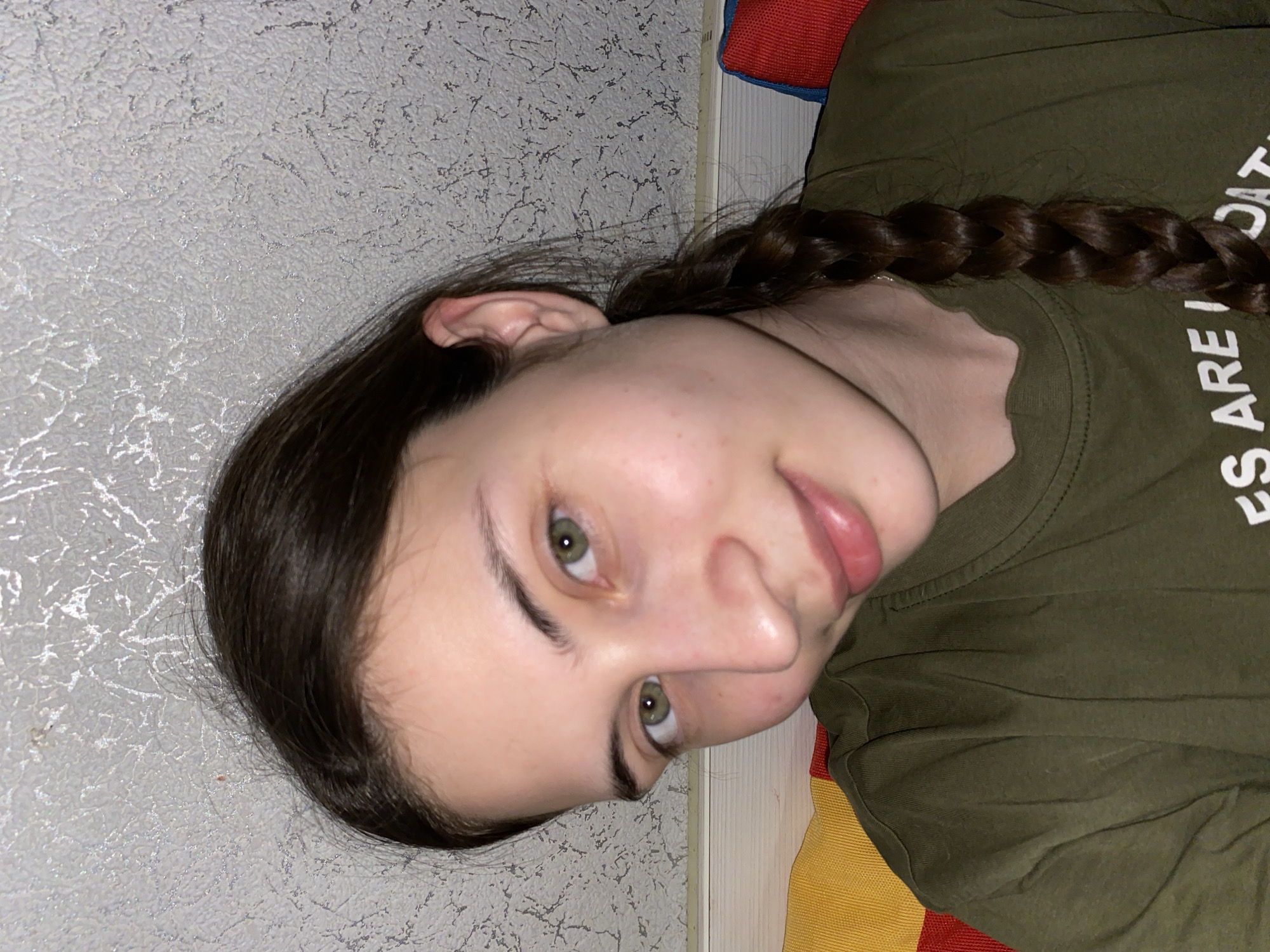 A young Caucasian woman with dark brown hair braided over her right shoulder is lying down. She is wearing an olive green t-shirt with white lettering partially visible. Her eyes are green. The background consists of a textured light gray wall. The bottom right corner shows a glimpse of multicolored fabric. There is no verifiable information to determine if this photo was taken in Votkinsk, Russia.