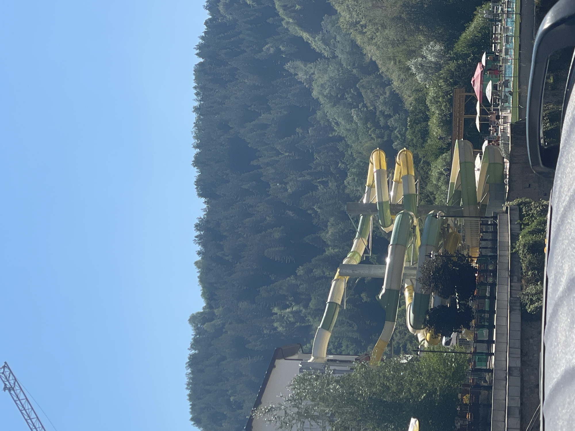 The image captures a water park situated in Banite, Bulgaria, with a dense, verdant forest covering a hillside in the background. Dominating the foreground are multiple colorful water slides, predominantly in yellow and green, curving and spiraling downwards. A building, possibly part of the resort or associated facilities, is visible on the left, partially obscured by trees. In the middle ground, a swimming pool area is set up with lounge chairs and umbrellas, some of which are red and white. Several individuals are present in the pool area, some near the slides and others relaxing by the pool. A construction crane is visible in the upper left corner of the image, suggesting ongoing development or maintenance in the vicinity. The sky above is clear and bright blue.