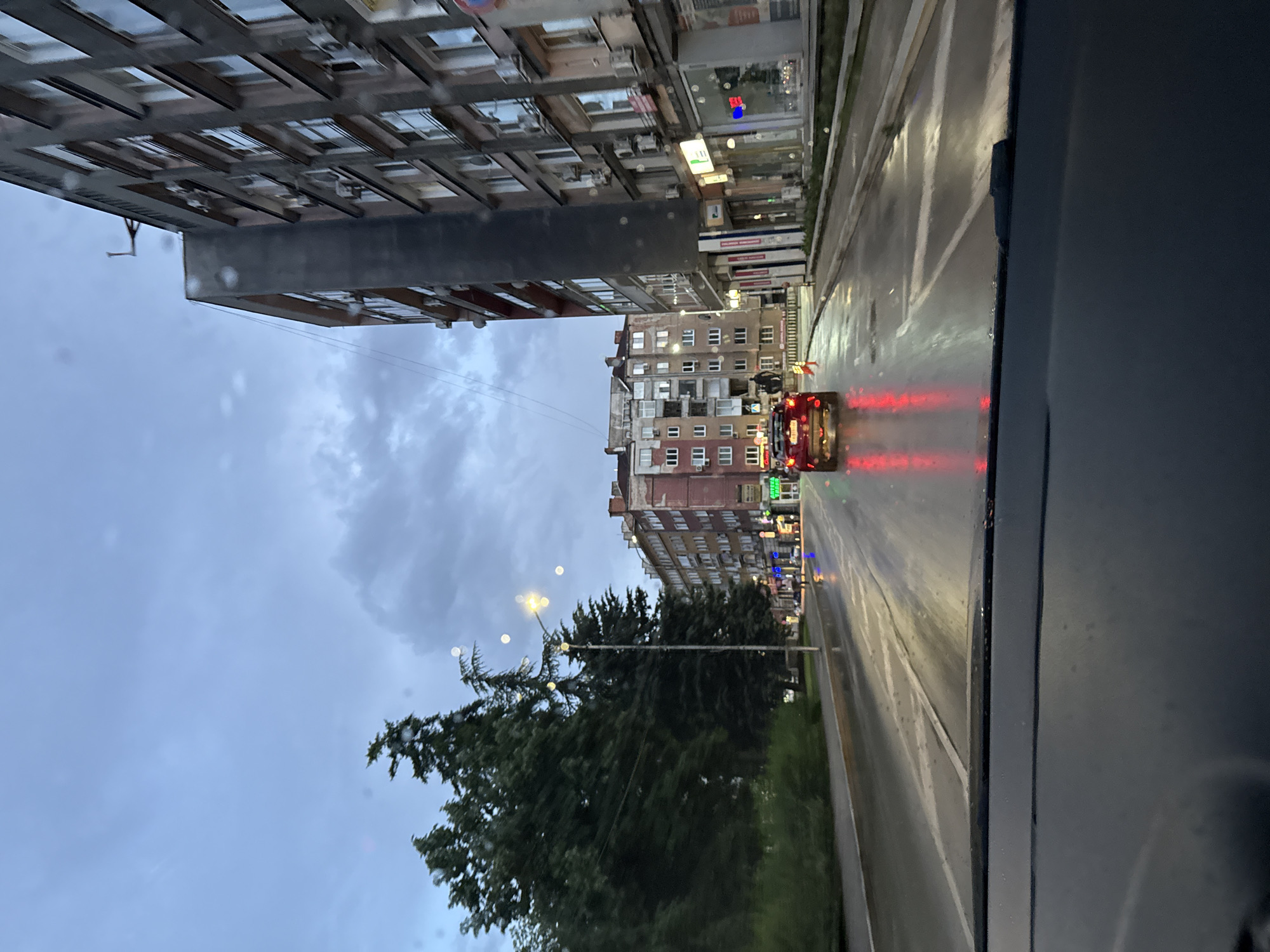 The image captures an urban street scene in Gabrovo, Bulgaria, likely during dusk or early evening, seen from inside a vehicle. The sky is overcast with light grey clouds, suggesting recent rain or impending precipitation, as the asphalt road is visibly wet and reflects streetlights and vehicle taillights.
On the left side of the frame, a row of multi-story buildings lines the street. The buildings feature numerous windows, some with balconies, and a mix of architectural styles, including older structures with exposed brickwork. One building appears more modern with uniform window frames and visible air conditioning units mounted on its exterior. Several commercial signs are affixed to the building facades.
The wet road occupies the middle ground, displaying white lane markings. A red car with brightly illuminated taillights is visible driving away from the viewer, its lights creating streaks of red on the slick surface.
To the right, a line of dense green trees, possibly conifers, runs alongside the road, interspersed with streetlights. The glowing streetlights create a hazy bokeh effect, potentially due to water droplets on the vehicle's window or the low light conditions. The far right edge of the image shows a dark section, indicating the interior frame of the vehicle from which the photograph was taken.
Visible text on a green sign on one of the buildings reads "OПТИКА" (Optics) with "T.M." below it. Another blue sign with partial text "TI." is also discernible. The overall atmosphere is dim and reflective, characteristic of a city street after rain.