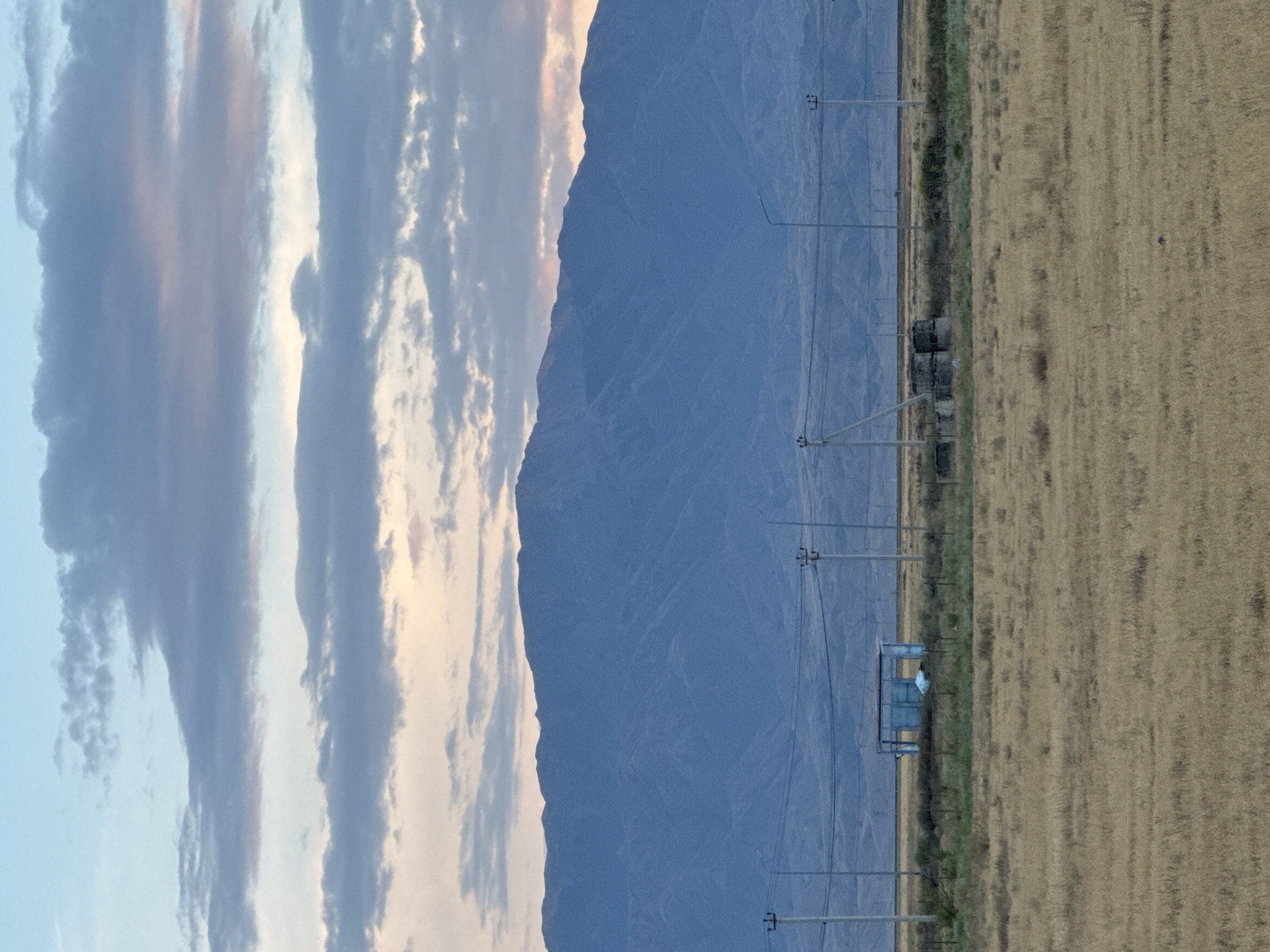 The image depicts a wide landscape under a cloudy sky, featuring a mountain range in the background. In the midground, there is a dry, grassy field with several utility poles and wires stretching across. A small, blue, open-sided structure with a white bag inside is visible in the field, as well as some concrete structures. The foreground is dominated by the dry grass. The scene appears to be in or near the city of Ақши, Kazakhstan.