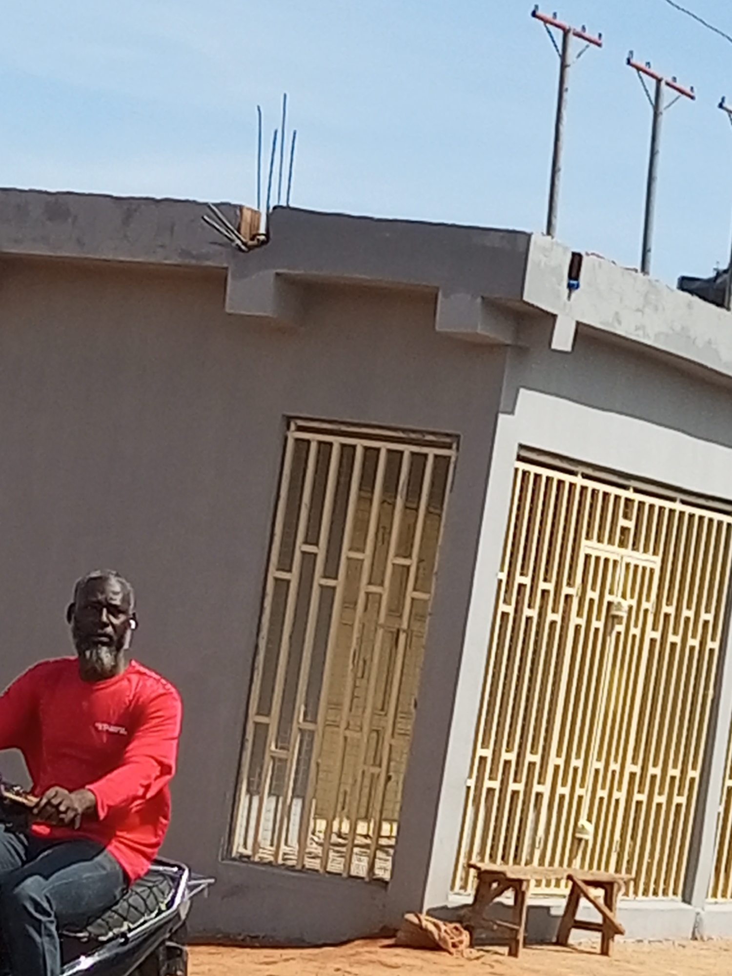 A man with a grey beard wearing a red long-sleeved shirt and blue jeans sits on a motorcycle in front of a grey building with barred windows and doors in Arawa, Nigeria. He is wearing white earbuds in both ears. The building appears to be under construction or recently completed, with rebar sticking out of the top. Two wooden stools and a burlap sack are placed on the sandy ground in front of the building. The sky is clear and blue, and two utility poles are visible in the background.