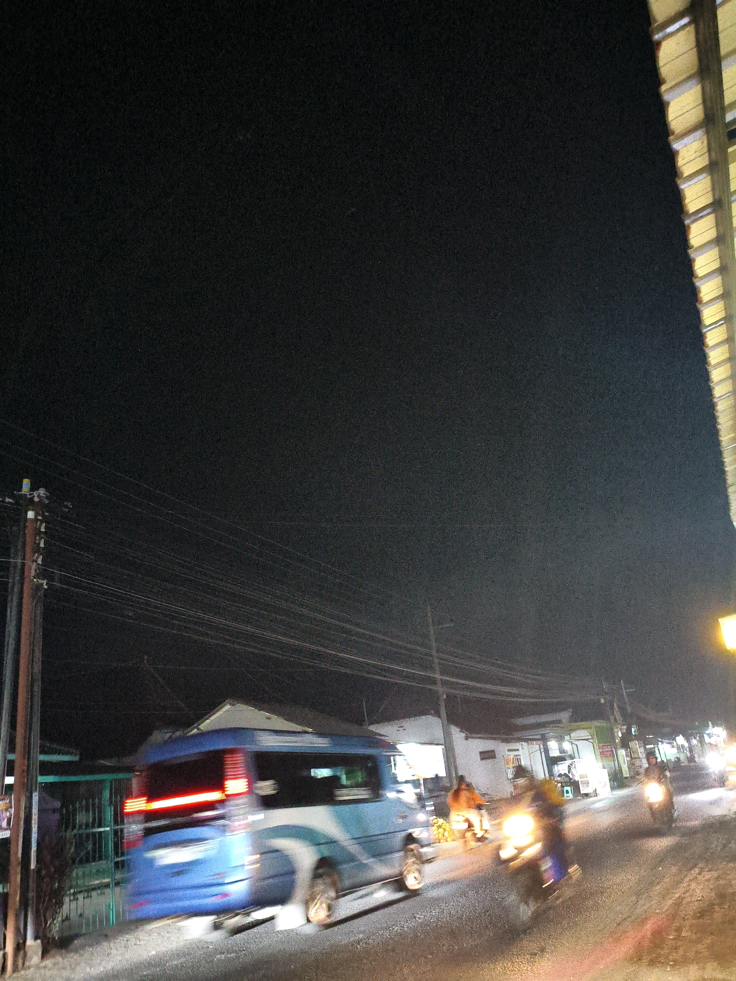 The image depicts a nighttime street scene in Tunggangri, Indonesia. A blue and white minivan is captured in motion on the left side of the frame, its taillights illuminated. Several motorcycles, also in motion, are visible further down the road, some with riders. Buildings line the street, some with illuminated storefronts or windows. Utility poles with numerous wires crisscross the sky. The road surface appears to be asphalt. The overall atmosphere suggests a bustling street at night.