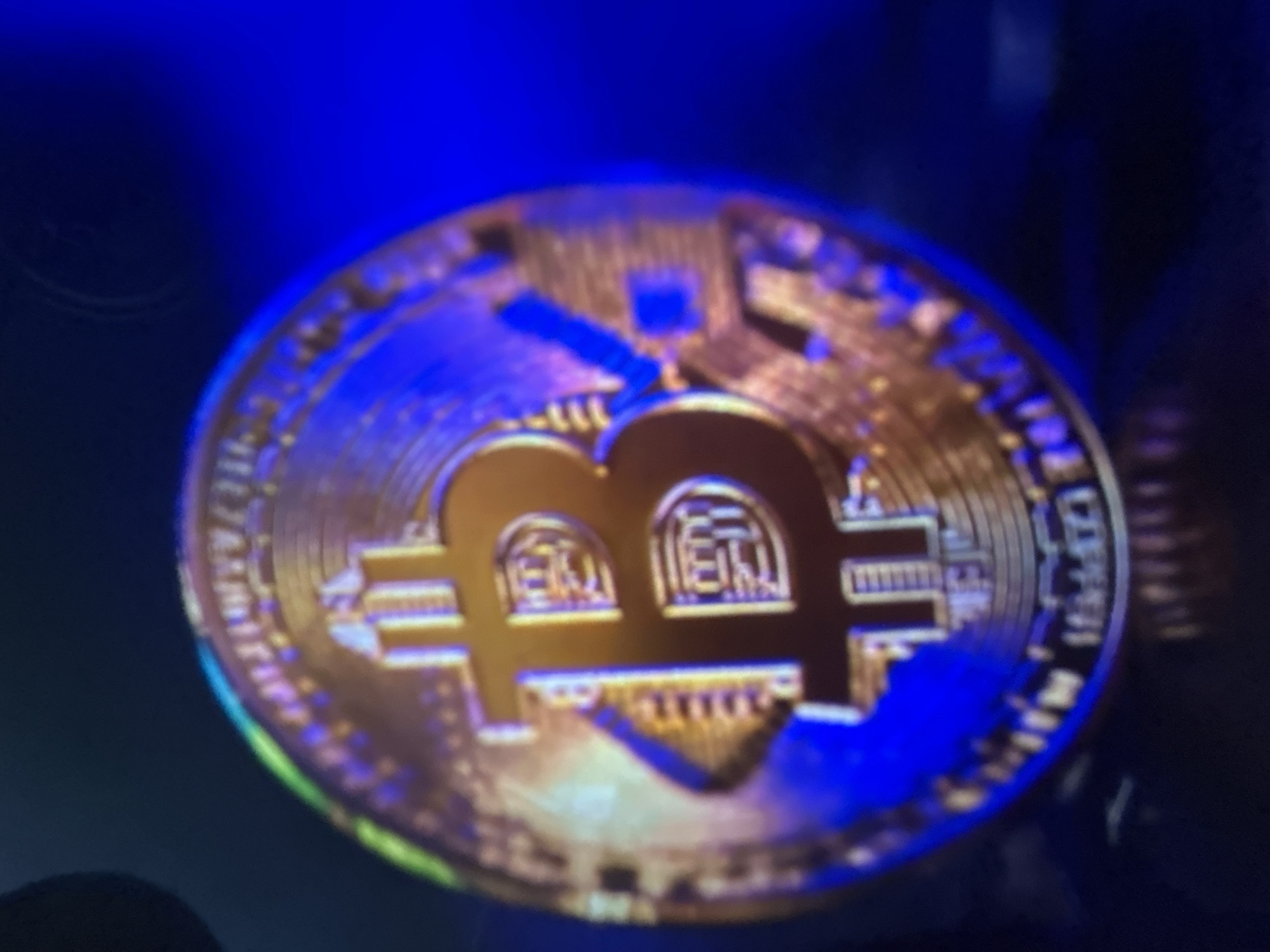 The media file is a close-up, blurry photograph of a golden Bitcoin coin. The coin is illuminated by blue light, which casts shadows and highlights on its surface. The background is dark and out of focus, with hints of blue and green hues, possibly from lighting or reflective surfaces.
The main subject is the Bitcoin coin itself, prominently displaying the characteristic "B" symbol with vertical lines. The coin's texture and intricate design are partially visible, though obscured by blurriness. There are no people or clear indications of a specific setting beyond the implied artificial lighting.
The image appears to be a still photograph, with no discernible activity or event taking place other than the presentation of the coin. The time of day, weather, and specific location cues within Gelendzhik, Russia, are not evident from the visual information provided. The image does not convey any specific emotions.
No readable text is visible on the coin or in the background due to the low resolution and blurriness. The focus is solely on the Bitcoin coin and the effect of the blue lighting.