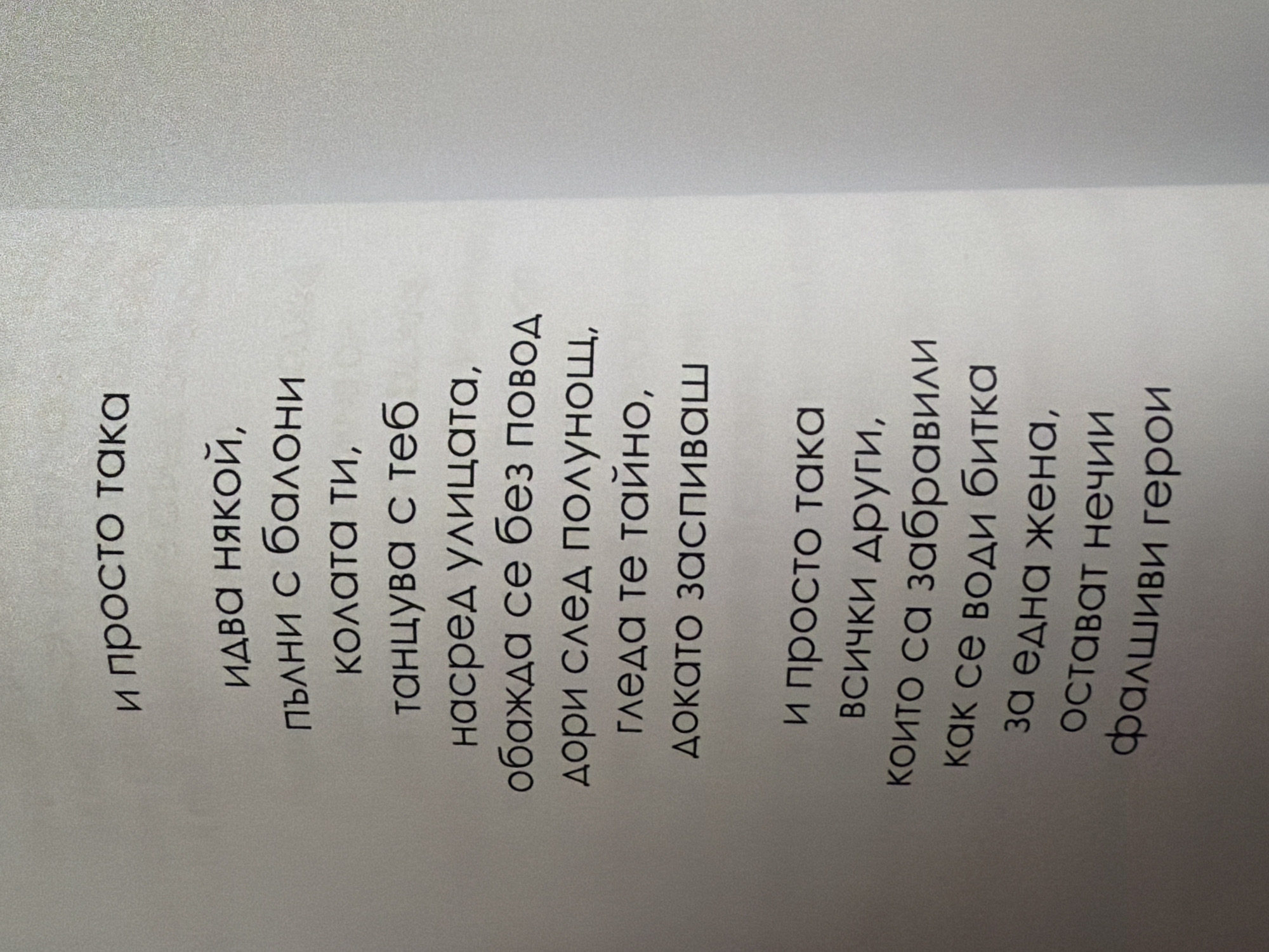 The image shows a page from a book with text written in Cyrillic. The text is a poem. The top of the page has the line "и просто така" (and just like that). Below that, it reads "идва някой, / пълни с балони / колата ти, / танцува с теб / насред улицата, / обажда се без повод / дори след полунощ, / гледа те тайно, / докато заспиваш" (someone comes, / filling your car with balloons, / dances with you / in the middle of the street, / calls without reason / even after midnight, / watches you secretly, / until you fall asleep). The bottom of the page has the line "и просто така" (and just like that) followed by "всички други, / които са забравили / как се води битка / за една жена, / остават нечии / фалшиви герои" (all others, / who have forgotten / how to fight a battle / for one woman, / remain someone's / fake heroes). The page is slightly off-center, with the majority of the text on the left side. The background is a light gray, textured surface, possibly a wall or paper. There is no indication of the city of Pazardzhik, Bulgaria, in the image.