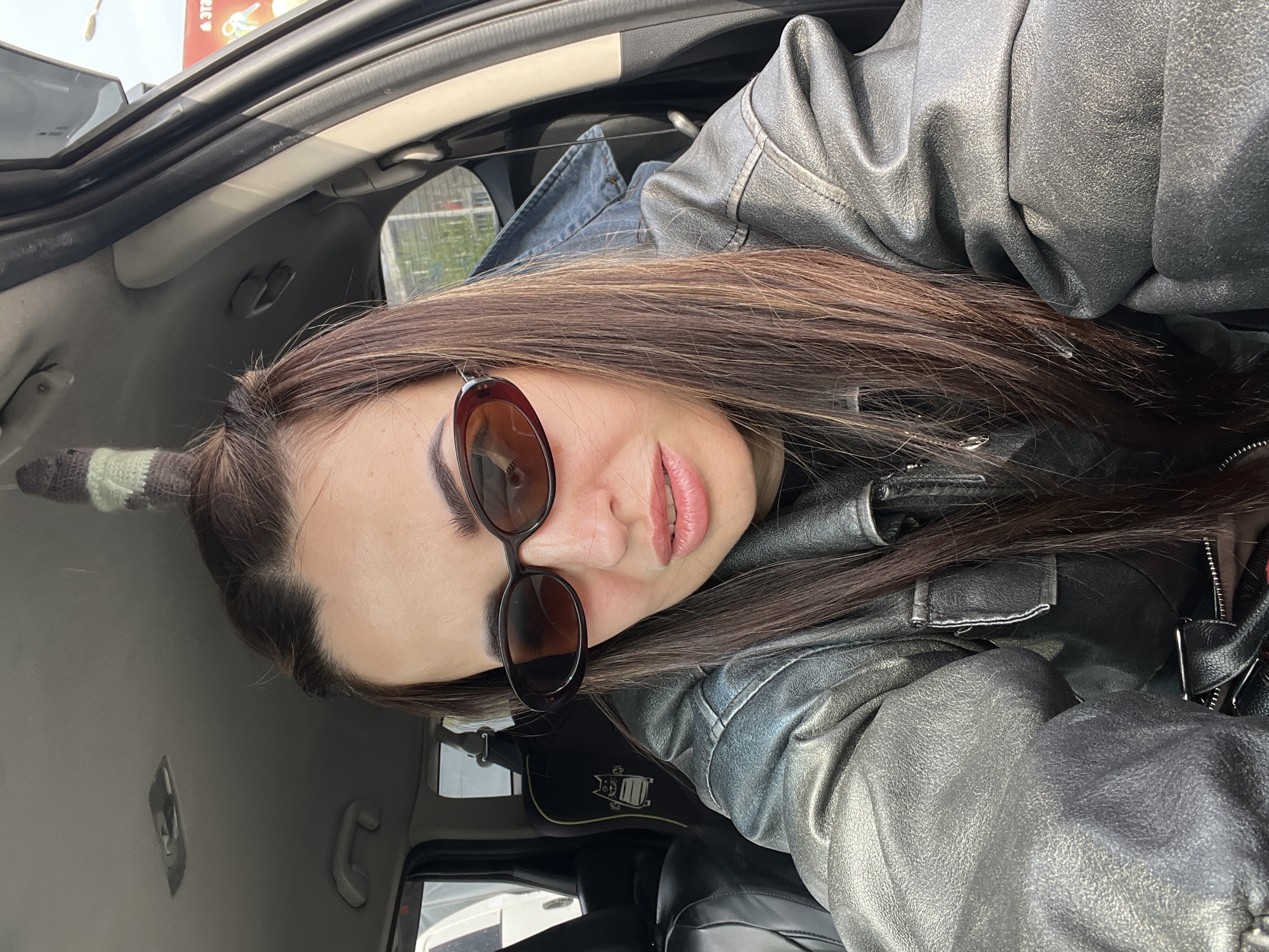 The image shows a woman with long brown hair wearing sunglasses and a black leather jacket. She is sitting inside a car with a gray interior. The location is likely an urban setting.