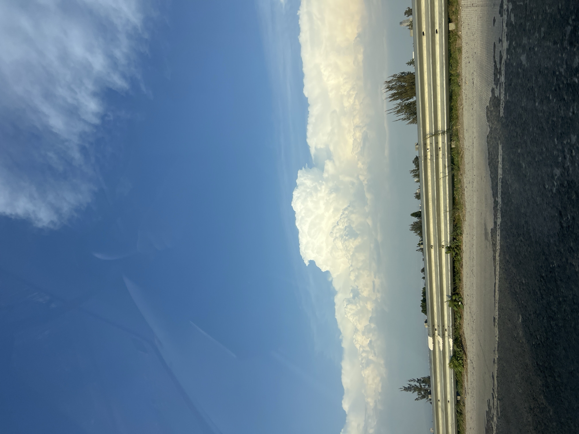 The image depicts a view from inside a vehicle, likely moving on a road. To the left, a clear blue sky dominates, with a few wispy clouds visible. To the right of the blue sky, there is a large bank of white, puffy clouds, seemingly towering and dense.
A highway guardrail runs along the right side of the frame, separating the road from the shoulder. Beyond the guardrail, there are glimpses of sparse vegetation, including what appears to be trees and shrubs. The road surface is visible in the foreground.
The image does not contain information to identify any city or location specifically. I cannot verify that the picture was taken in San Bernardino Tlaxcalancingo, Mexico.