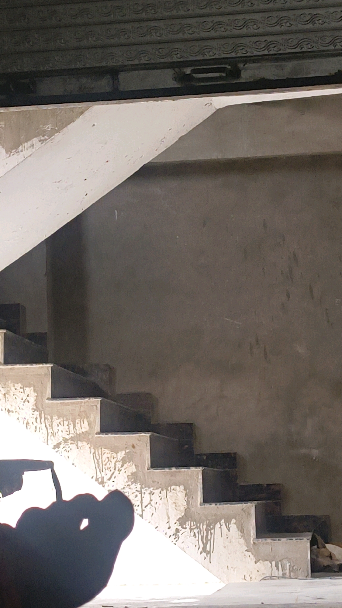 The image depicts an unfinished staircase, likely under construction, in Manjhanpur, India. The steps are partially completed, with some sections appearing to be concrete and others covered in what looks like plaster or a rough finish. There are drips and stains on the stairs, suggesting recent work or exposure to the elements. A shadowy silhouette of what appears to be a person's hand or foot is visible in the lower left corner, casting a distinct shadow on the staircase and the surrounding wall. The wall behind the stairs is a neutral, gray-brown color, also appearing unfinished. Above the staircase, a dark, textured structure is visible, possibly a ceiling or a decorative element. The overall scene conveys a sense of ongoing construction and a raw, unrefined environment.