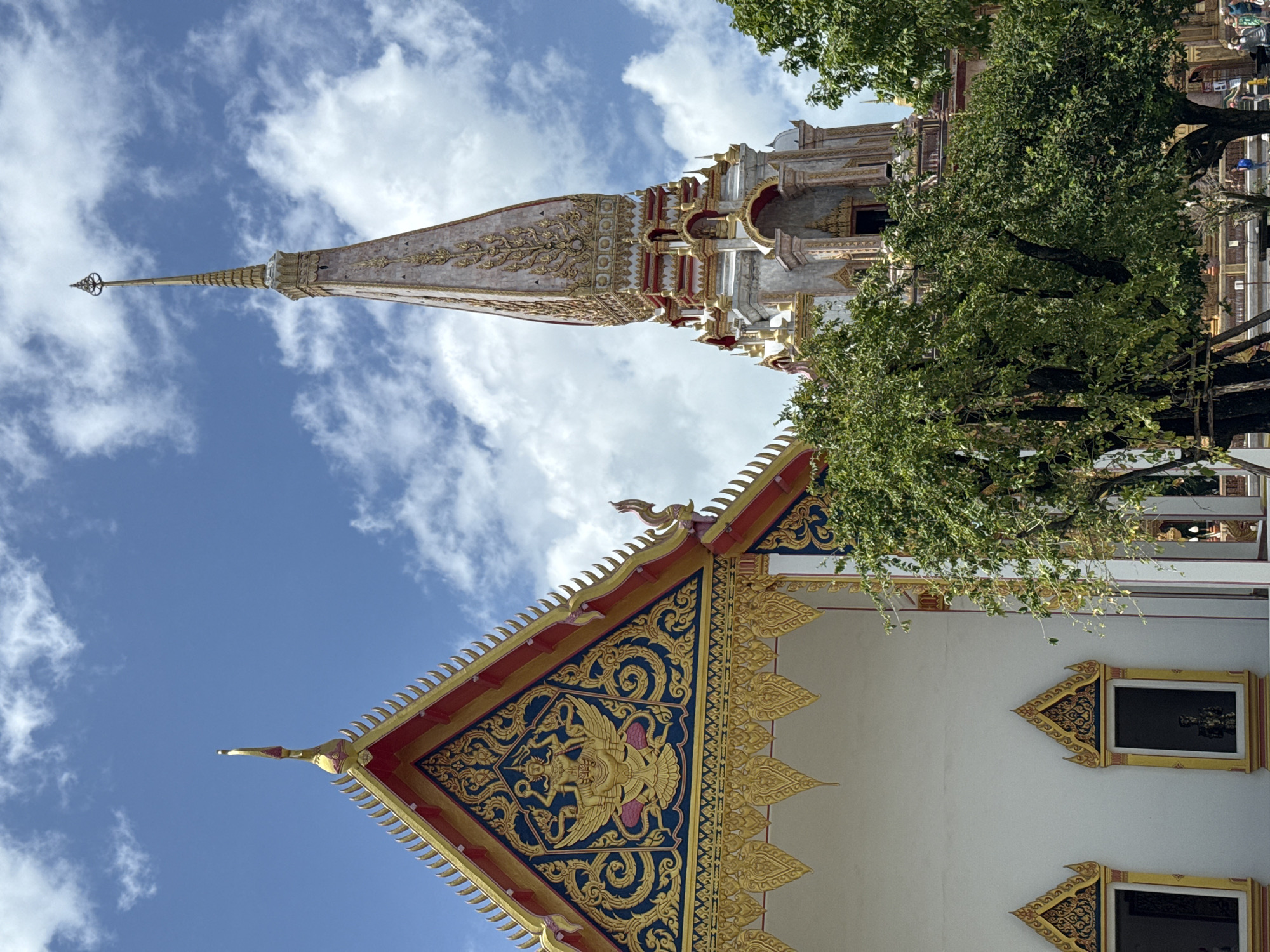 This is a medium-shot photograph taken outdoors in Chalong, Thailand, focusing on a Buddhist temple. The sky is blue with scattered white clouds. In the foreground, the white wall of a temple building is visible, adorned with intricate gold and dark blue decorative elements on its triangular gable. Two windows with similar decorative framing are set into the wall, with faint statues visible inside. To the right, lush green trees partially obscure a tall, ornate stupa, which is a dominant feature of the temple's architecture, featuring intricate gold embellishments against a pale, textured background, with red and gold trim around its lower sections. The overall scene depicts a serene and architecturally rich religious site on a bright, clear day.