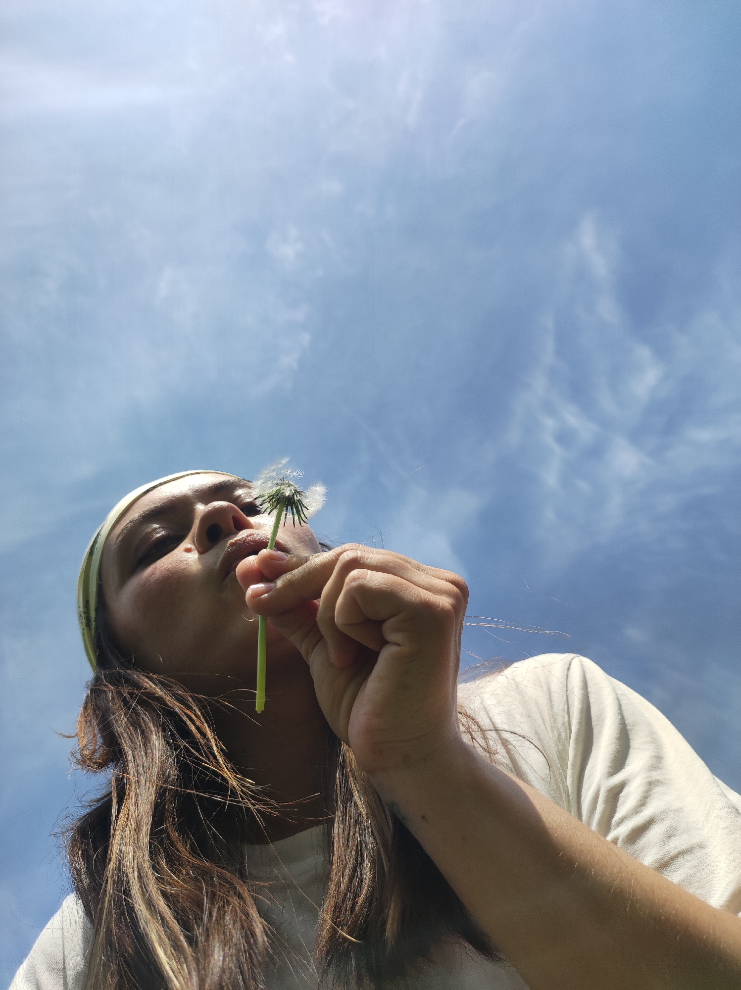 A low-angle shot depicts a person blowing on a dandelion held between their fingers. The subject is a young individual with long, light-brown hair and a light complexion, wearing a white t-shirt and a pale green headband. They are positioned against a blue sky dotted with wispy clouds. The dandelion's seed head is partially dispersed. No features indicate the location is specifically in the city of Фоки, Russia.