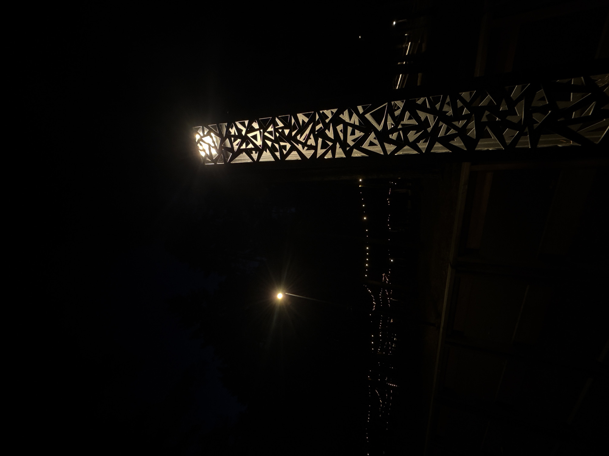 The image captures a nighttime or very dark scene, dominated by artificial lighting. The primary subject is a prominent horizontal light fixture positioned in the upper portion of the frame. This fixture is a long, rectangular beam with an intricate, laser-cut pattern of varied geometric triangles. Light emanates brightly from within, illuminating the metallic or opaque surfaces of these triangular cutouts and creating a distinctive visual texture.
To the left and slightly below the main fixture, a secondary, smaller light source is visible, appearing as a bright, star-like point with a noticeable flare. Further to the right, a vertical element containing a string of numerous small, sparkling lights descends into the darkness, suggesting a decorative light installation or a structural feature adorned with lights.
The overall setting is characterized by profound darkness, making it difficult to discern specific location details beyond the illuminated elements. The scene is devoid of people or any discernible activity, placing the sole emphasis on the modern and abstract lighting design. The context suggests this could be an architectural or artistic light installation in Votkinsk, Russia. There is no visible text in the media file.