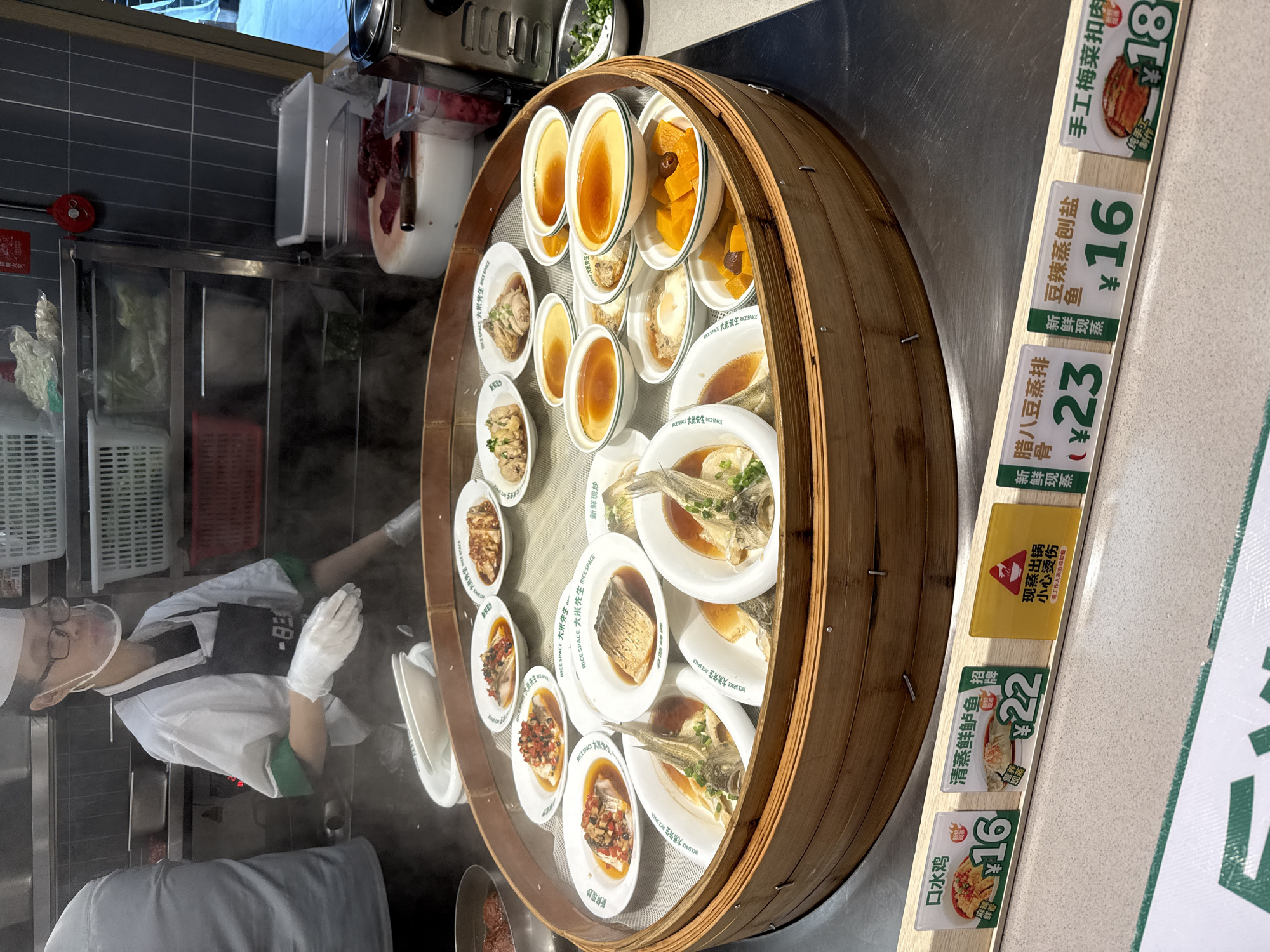 The image depicts a food stall in Tianhe District, China, showcasing a variety of steamed dishes. A food handler, wearing a white uniform, black apron, clear face shield, and white gloves, is visible in the background, tending to the food. The main subject is a large, circular bamboo steamer filled with numerous small plates of food.
The steamer contains an assortment of dishes, including several whole steamed fish, some topped with chili and herbs, and others with a sauce. There are also plates of what appear to be steamed chicken, steamed eggs (custard), diced pumpkin, and possibly some sort of meat dish. The food is arranged in concentric circles within the steamer.
In the foreground, there are price tags for some of the dishes. One tag shows "口水鸡" (Sichuan Boiled Chicken) for ¥16. Another tag displays "清蒸鲜鲈鱼" (Steamed Perch) for ¥22. A yellow tag with a warning symbol and text indicating "小心烫" (Be careful of the heat) is also present. Further to the right, other price tags are visible, including "腊八豆蒸排骨" (Laba Bean Steamed Pork Ribs) for ¥23, "豆辣蒸创鱼" (Spicy Bean Steamed Fish) for ¥16, and "手工梅菜扣肉" (Handmade Preserved Vegetable Pork Belly) for ¥18.
The environment appears to be an indoor food court or market, with stainless steel surfaces and tiled walls visible in the background. Steam is rising from the food, indicating it is freshly prepared. The lighting is bright, highlighting the food and the details of the scene.