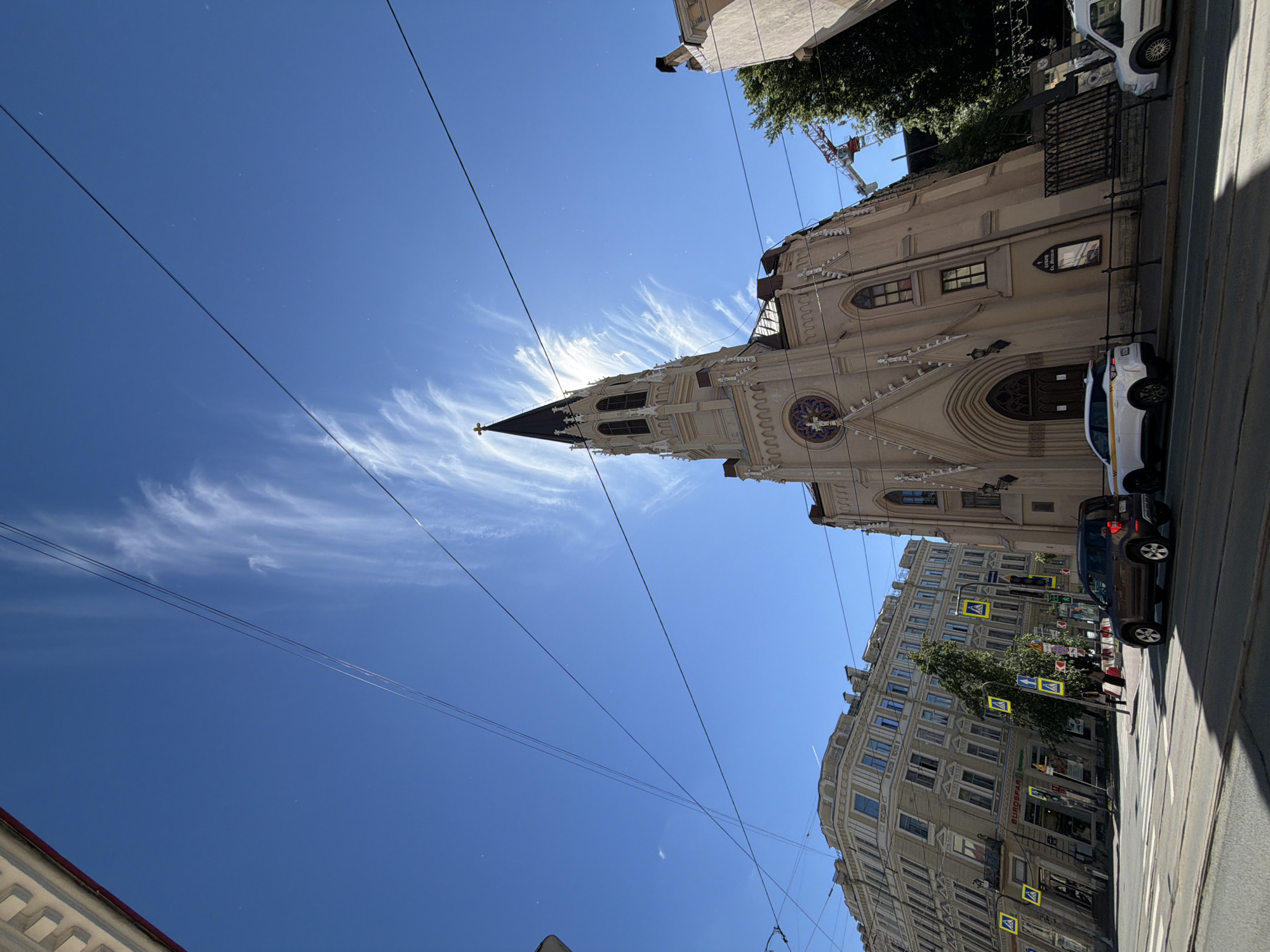 The image captures a low-angle view of a neo-Gothic style church with a tall, pointed spire, prominently featured against a bright blue sky with wispy white clouds. The church building is a light beige color, with arched windows and intricate architectural details. A cross tops the spire, and the sun is positioned behind it, creating a lens flare effect and obscuring the very top of the spire.
In the foreground, a street with tram tracks runs horizontally. Two cars are visible on the street: a dark brown SUV and a white car. To the left of the church, a multi-story building with a European architectural style is visible. This building has many windows and displays yellow pedestrian crossing signs at its base. The street is lined with trees and streetlights.
Further to the right, a construction crane is visible in the background, suggesting ongoing development or renovation in the area. The overall scene is a city street in Saint Petersburg, Russia, likely on a clear day. The image's perspective is wide, emphasizing the sky and the grandeur of the church.