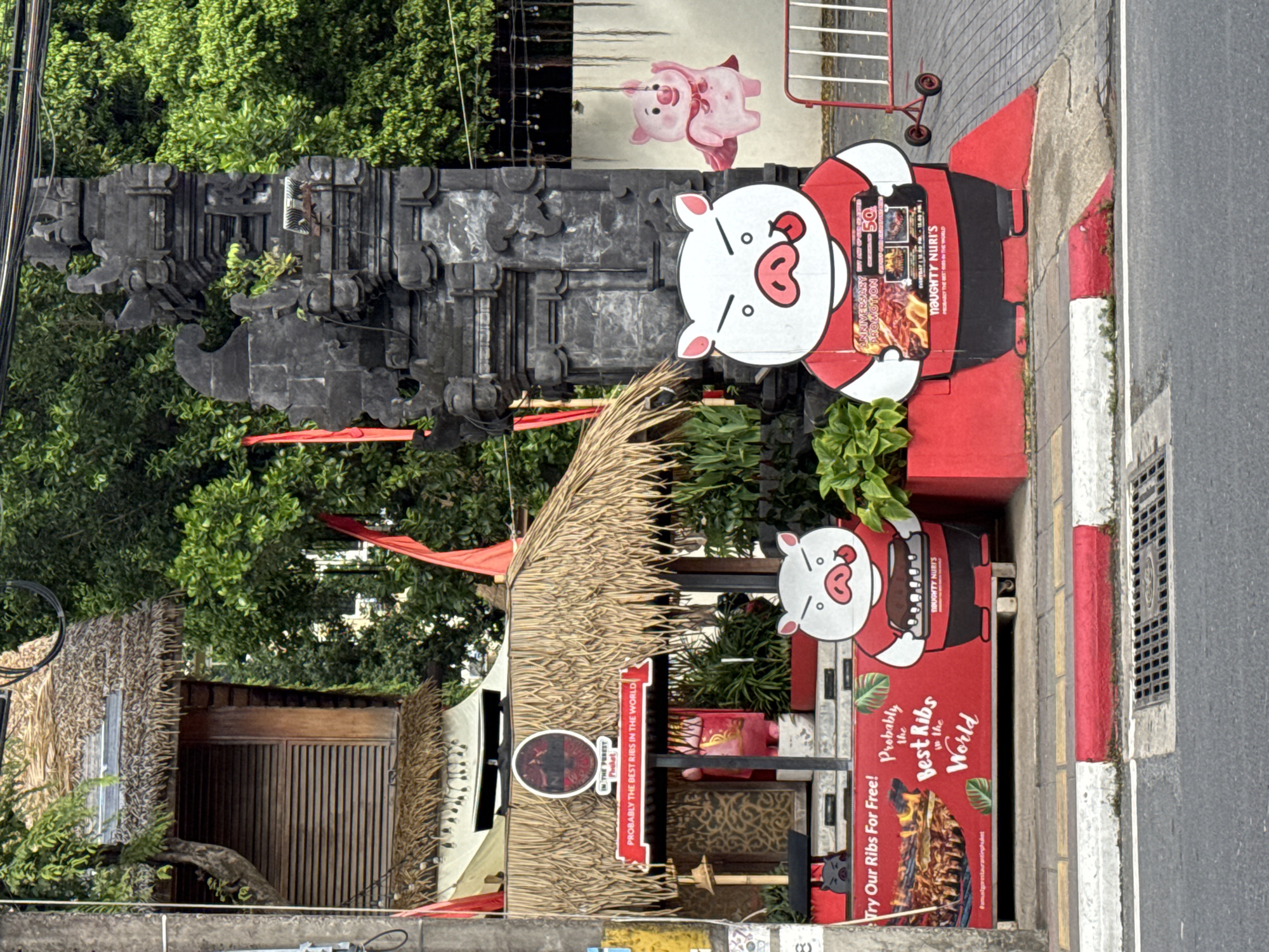 An outdoor scene in Patong, Thailand features a roadside eatery with a thatched roof and wooden shutters. Two large, cartoonish pig figures stand outside, one holding a tray of ribs and another with a cape. A pink pig illustration hangs from a wire above them. The eatery has a red banner proclaiming "Probably the Best Ribs in the World." In the background, a stone archway and lush greenery are visible. The street is paved with concrete and bordered by a red and white curb. A metal barrier with wheels is placed on the right side of the frame.