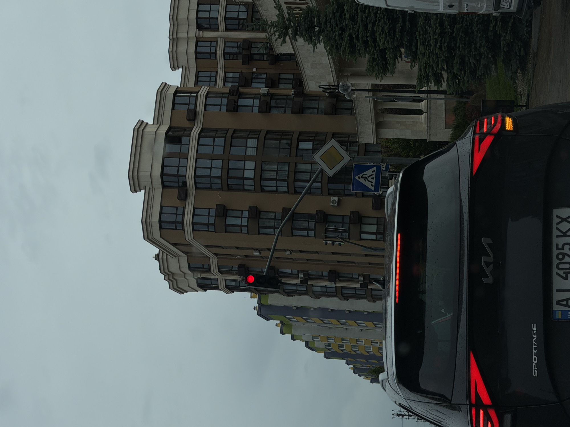 The image, taken in Sofiivska Borshchahivka, Ukraine, captures a street scene on an overcast day, suggesting gloomy or rainy weather conditions, as evidenced by the wet surface of the car. The foreground is dominated by the rear of a black Kia Sportage SUV, seen from the right side of the frame. Its distinctive red brake lights are illuminated, and the right turn signal flashes amber. The car's rear displays "KIA" and "SPORTAGE" emblems, along with a Ukrainian license plate reading "AI 4095 KX" and the "UA" country identifier.
In the mid-ground, a red traffic light is visible, indicating that the vehicle is stopped. Nearby, two road signs are mounted on poles: a yellow diamond-shaped priority road sign and a blue square sign indicating a pedestrian crossing.
The background features several multi-story buildings. A prominent modern building with a beige/light brown facade and numerous dark windows dominates the center, showcasing architectural details and several visible air conditioning units. Behind this, another multi-story residential building with a more colorful facade, featuring segments of yellow, green, and blue, is partially visible. Dense green foliage from trees adds a natural touch to the urban landscape. The sky is a uniform gray, reinforcing the overcast weather. No people are visible in the scene.