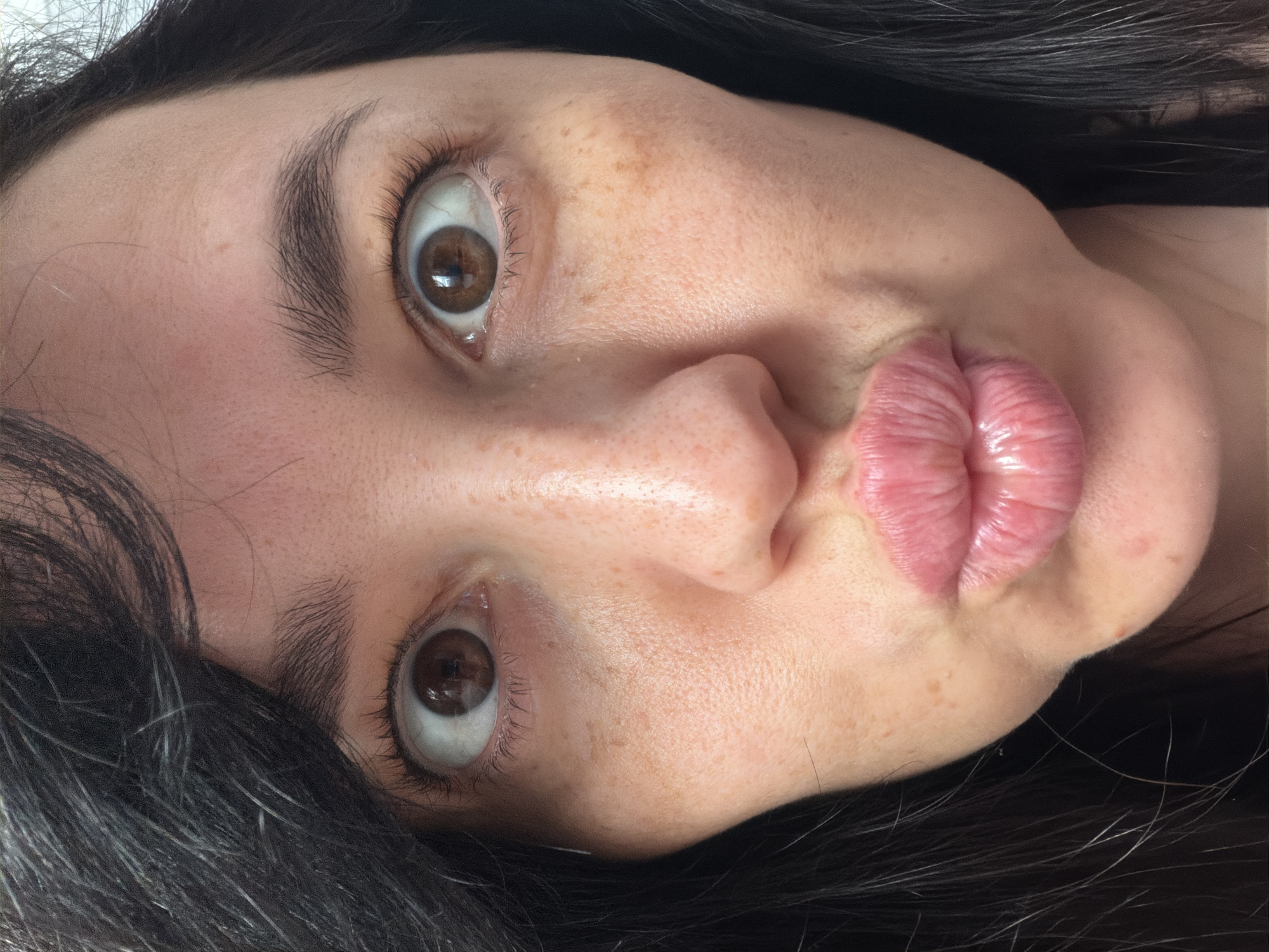 A close-up, head-on, chest-up shot captures the face of a young adult woman. Her dark hair frames her face, and her eyes are wide open and looking directly at the camera. Her eyebrows are dark and well-defined. She is puckering her lips, which are full and glossy pink. The woman's skin has a light complexion with visible freckles and some slight redness. The background is a plain, light-colored surface, out of focus. There is no discernible location in Moscow, Russia, or any other identifiable environmental features in the image.